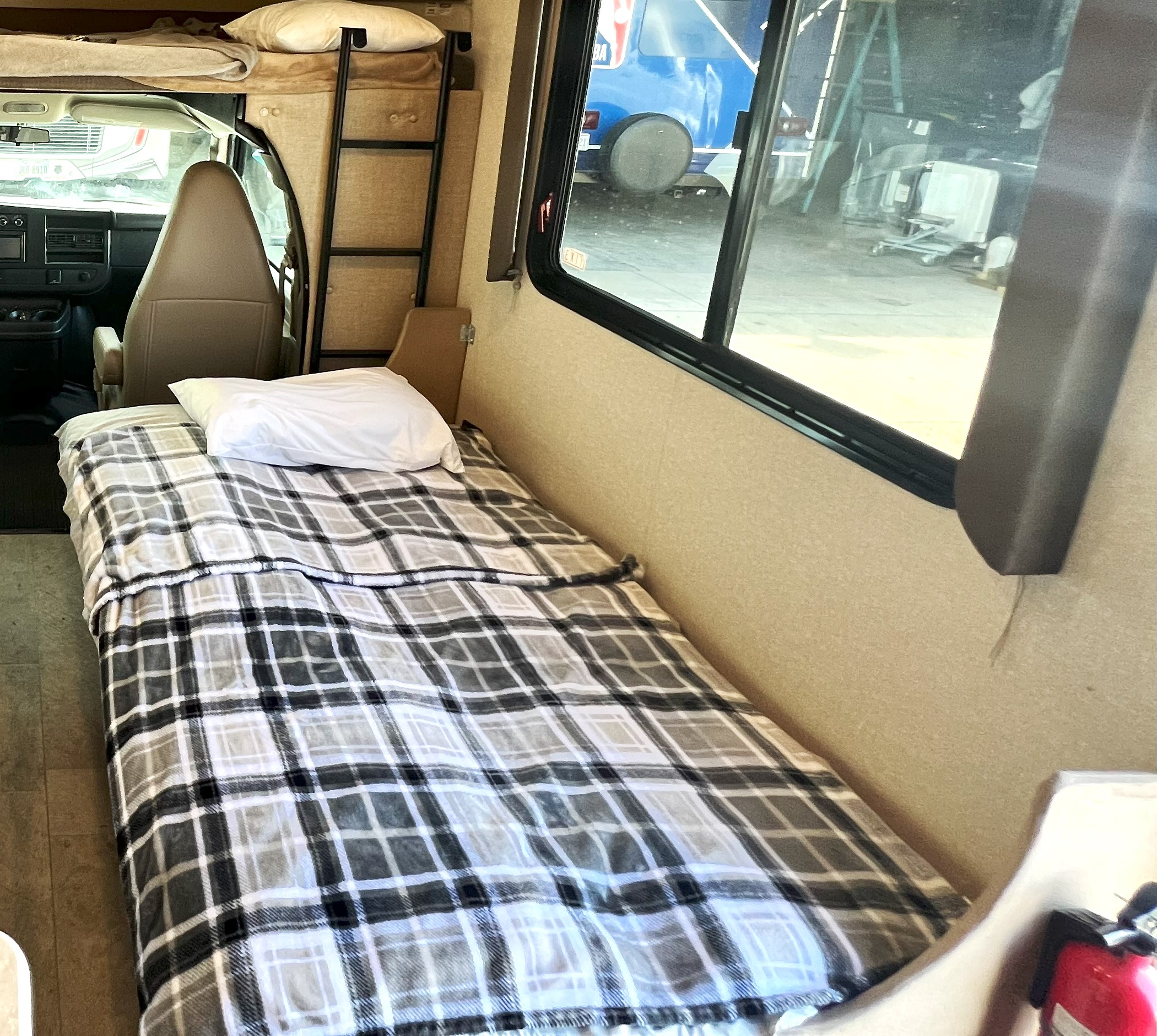 Inside an rv rental: a cozy single bed with a plaid blanket, driver's cab, and ladder to an overhead bunk.. Coachman Freelander QB 2019