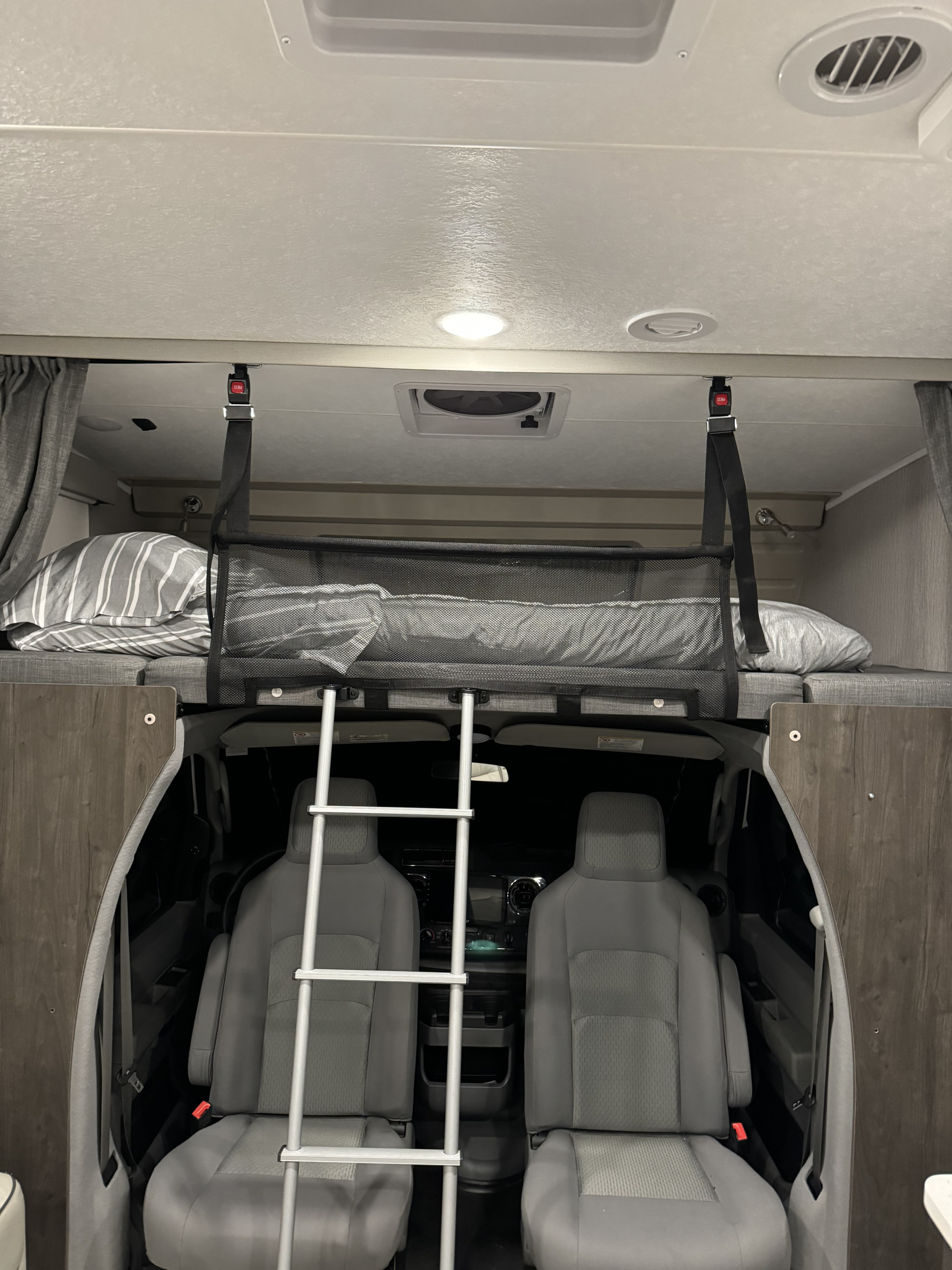 Inside an RV rental: a spacious over-cab bunk bed with safety net and ladder, viewing the front cockpit and grey seats.. Forest River Coachmen Leprechaun Premier 2022