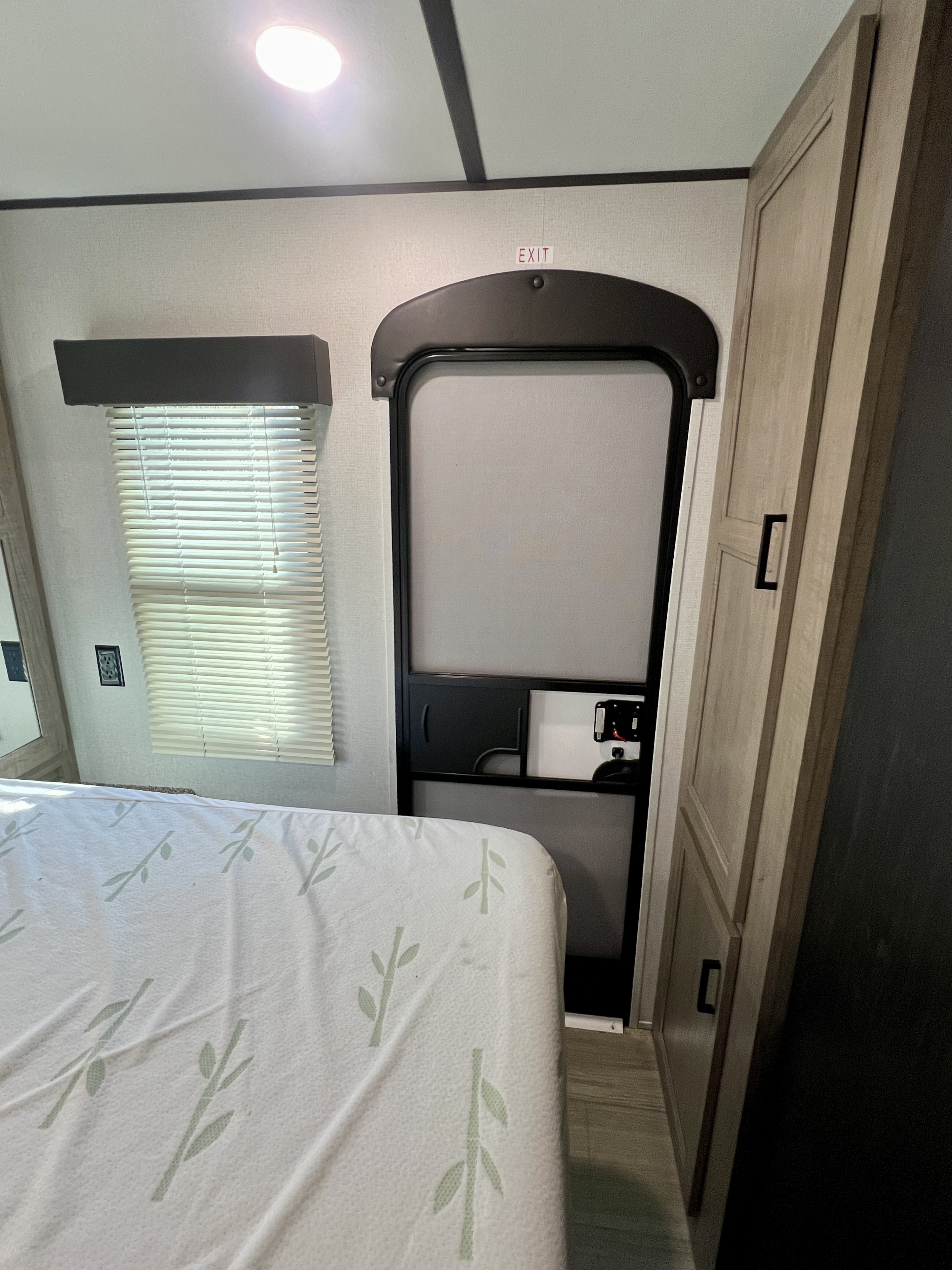 Interior of an RV rental: a bright, compact bedroom featuring a bed, window with blinds, and the main exit door.. Keystone Hideout 2020