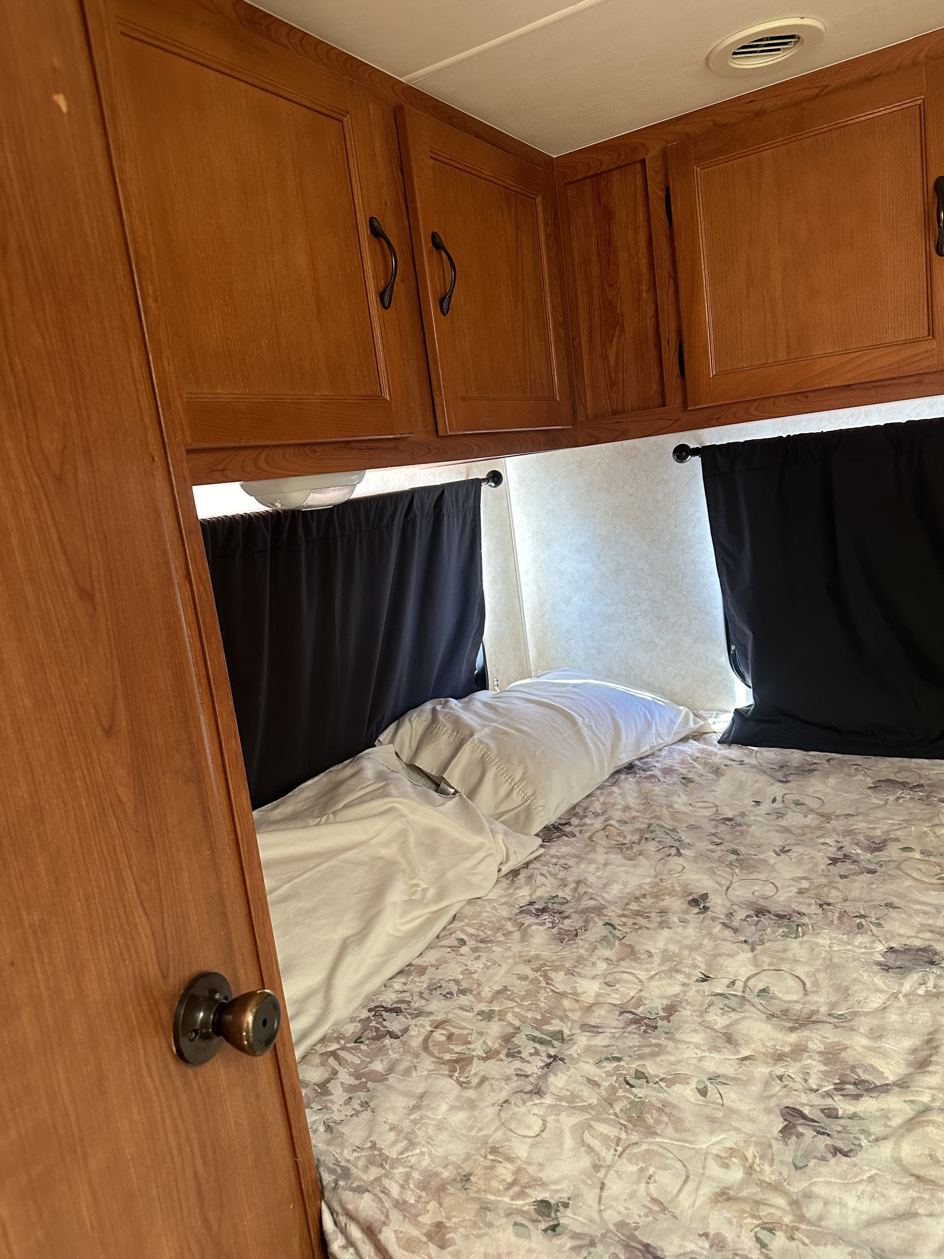 Cozy RV rental bedroom features wood cabinets, floral bed, black curtains, and a side door.. Coach House Freelander 2011