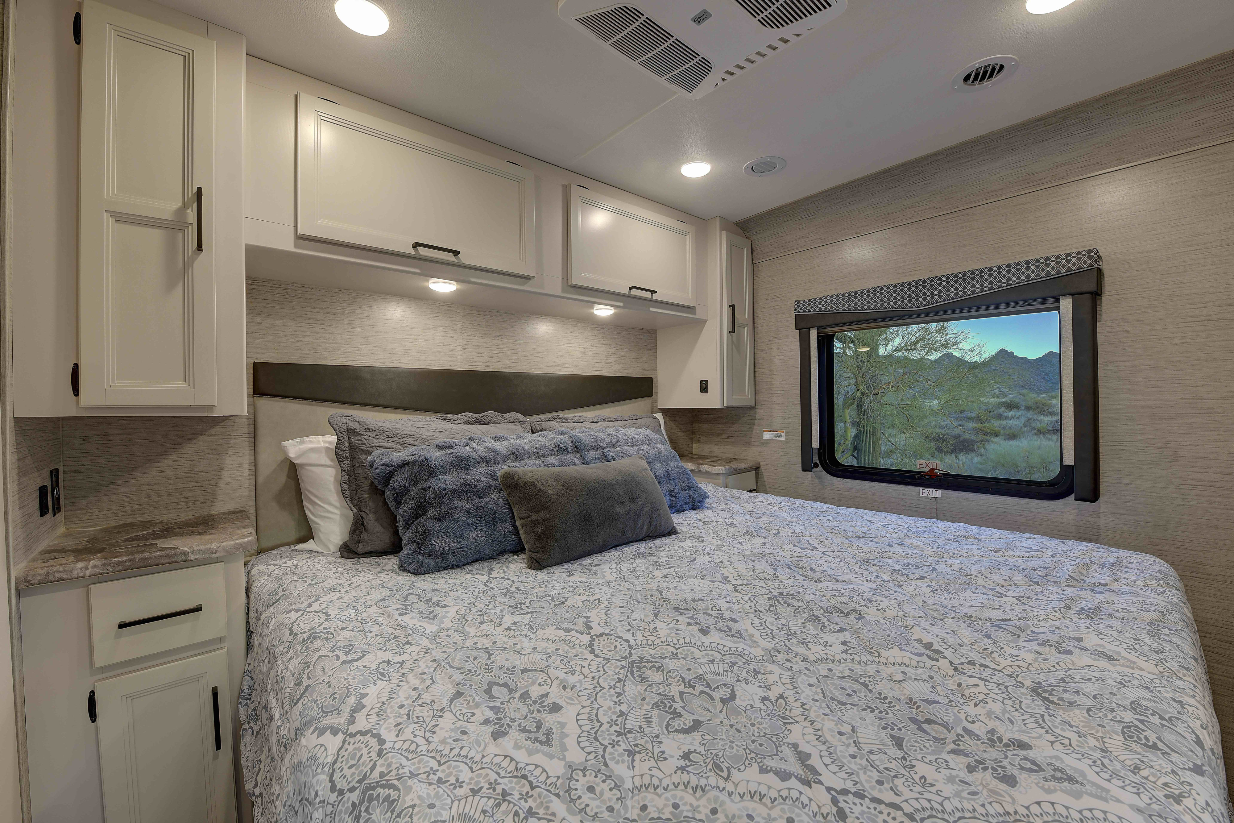 Cozy RV rental bedroom features a comfortable bed, overhead storage cabinets, and a scenic desert view through the window.. Thor A.C.E. 2022
