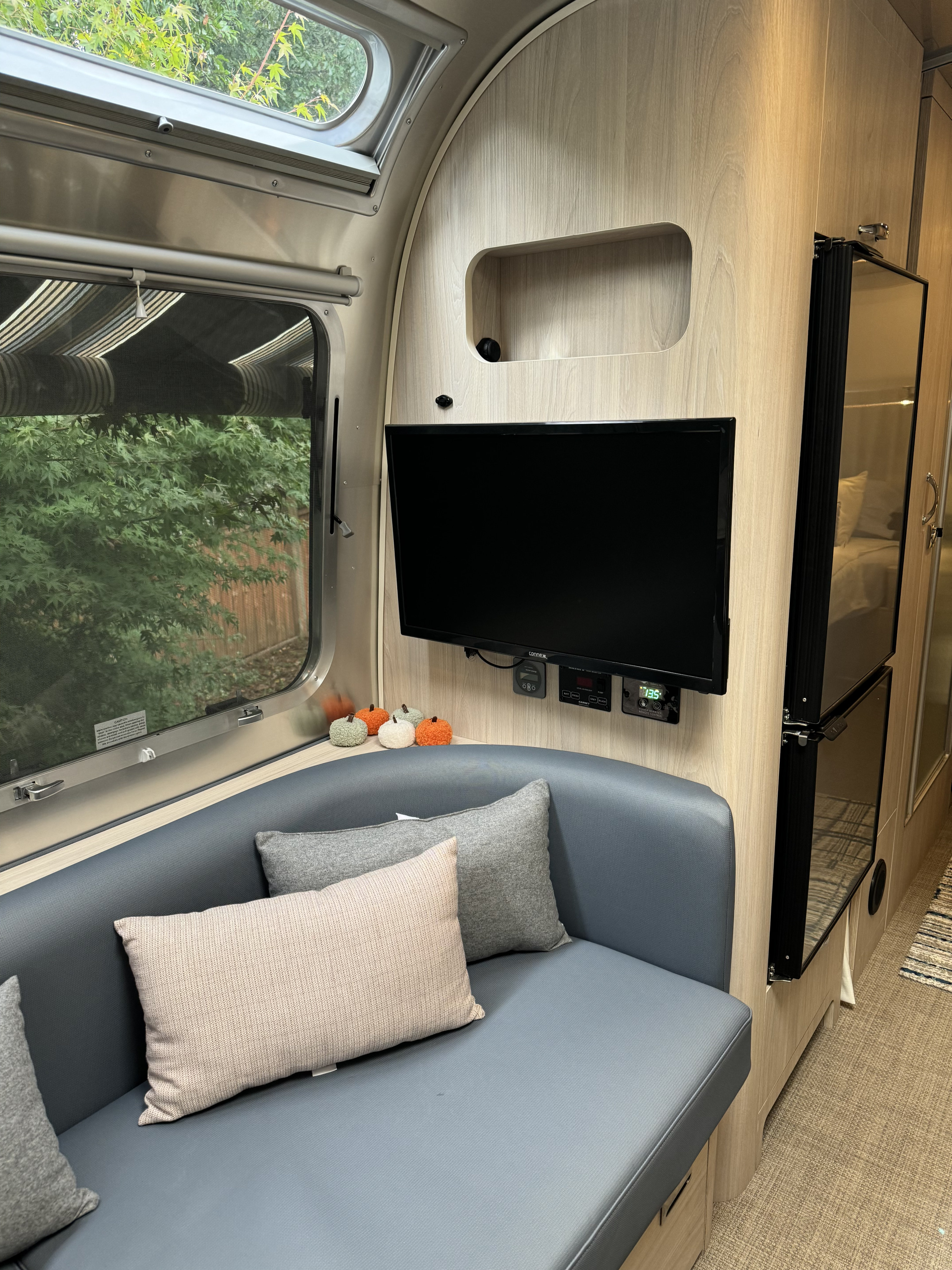 Cozy RV rental interior: A blue sofa, mounted TV, window with tree view, & fridge glimpse.. Airstream Globetrotter 2024