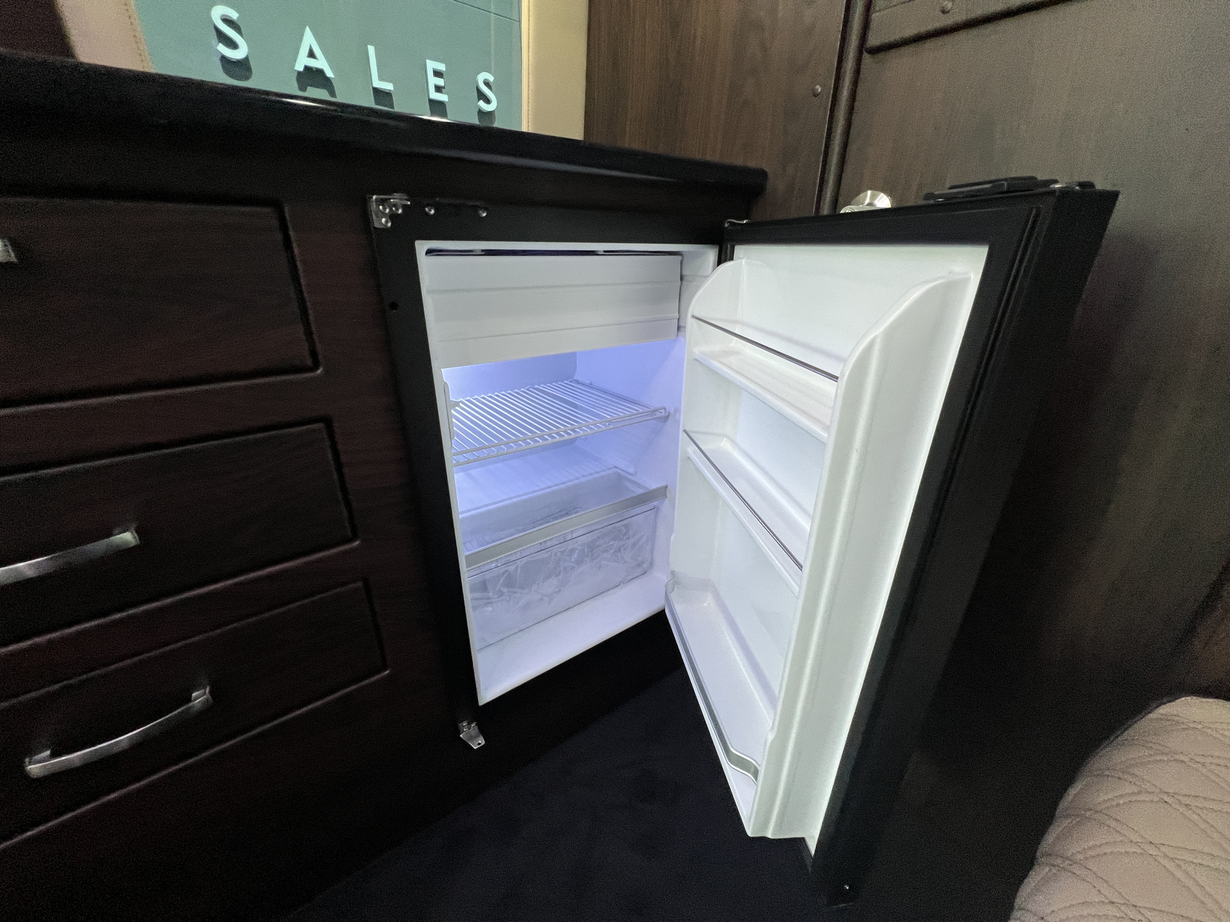 Inside an RV rental: a bright, open mini-fridge built into dark cabinetry alongside storage drawers.. Midwest Automotive Designs Day Cruiser 2024