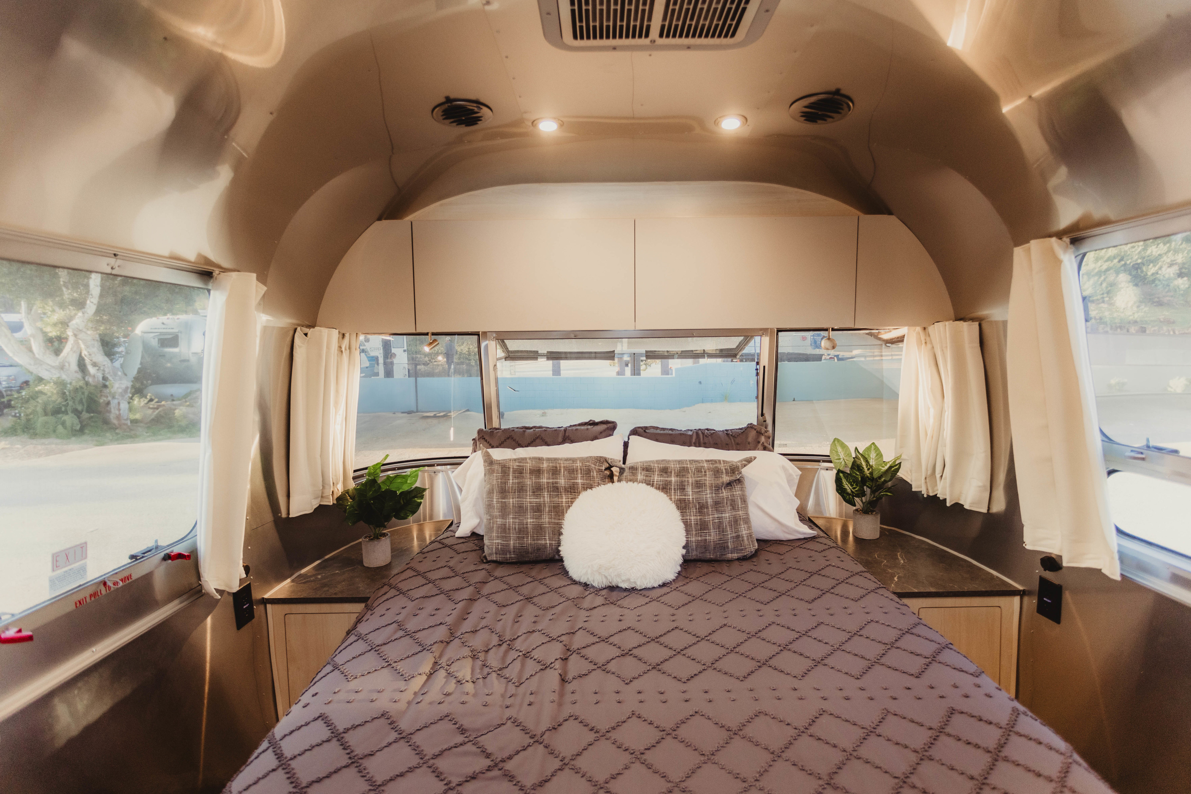 This RV rental interior shows a cozy bedroom with a patterned bed, pillows, and bright windows with white curtains.. Airstream Flying Cloud 2024