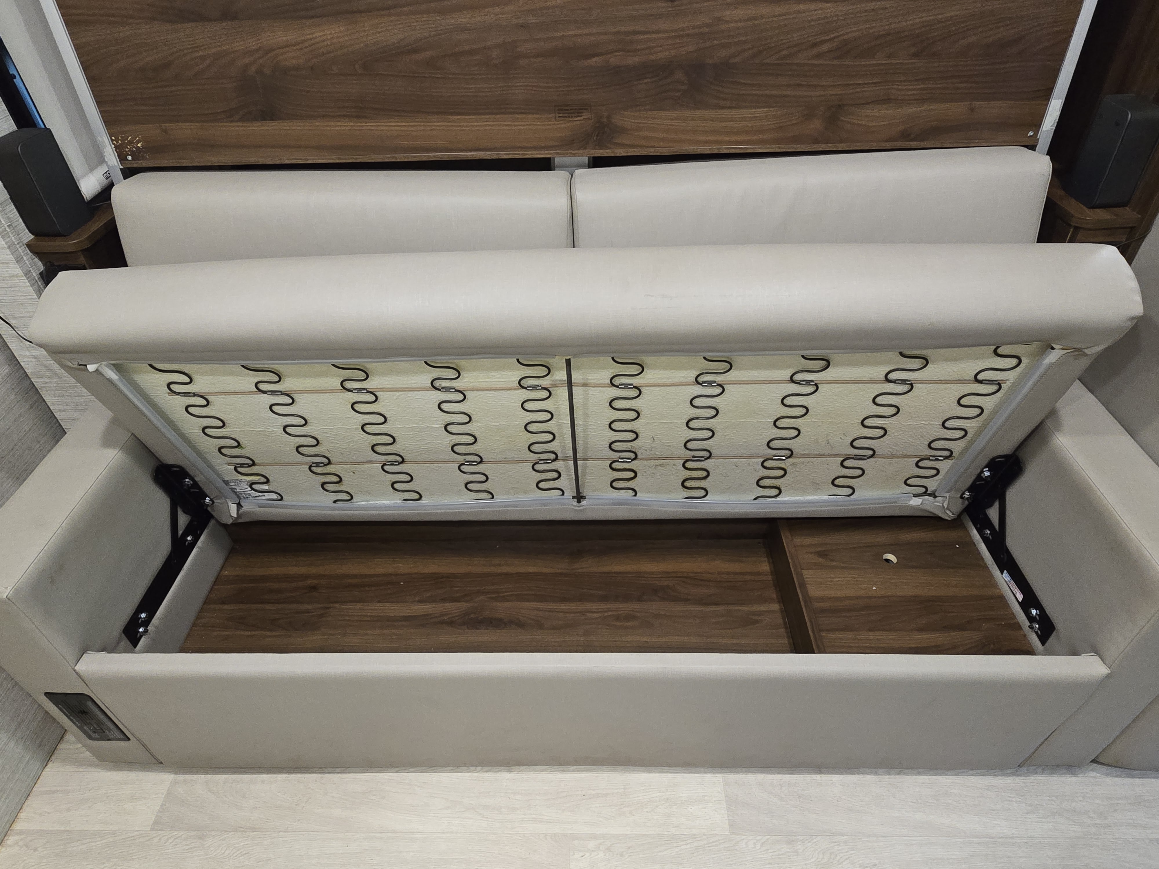 Inside an RV rental, a light-colored sofa cushion is lifted, exposing metal zigzag springs above a dark wood storage area.. Winnebago View 2022