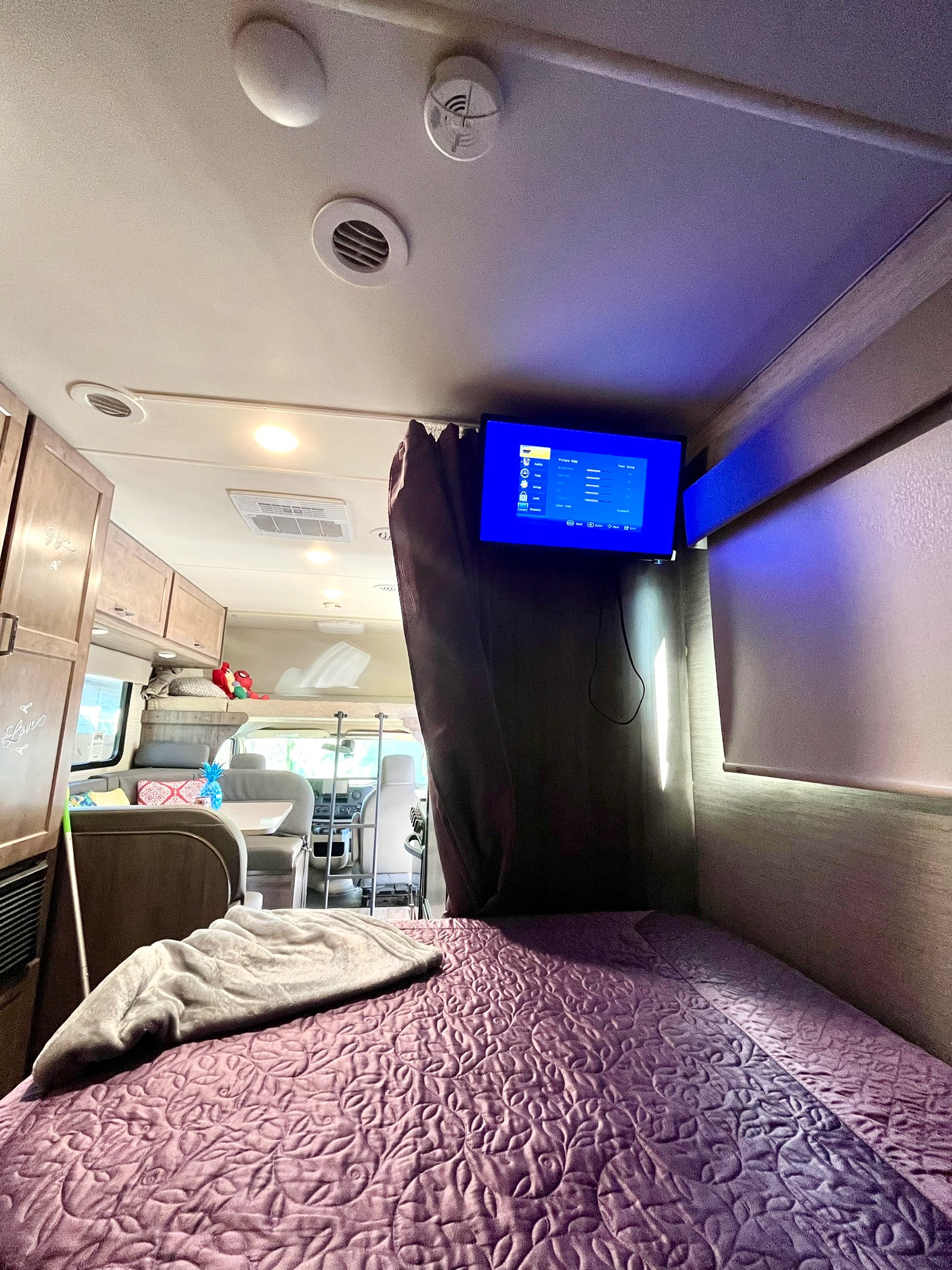Inside an RV rental: a comfortable bedroom with a purple quilted bedspread and mounted TV, extending to a living area.. Winnebago Minnie Winnie 2021