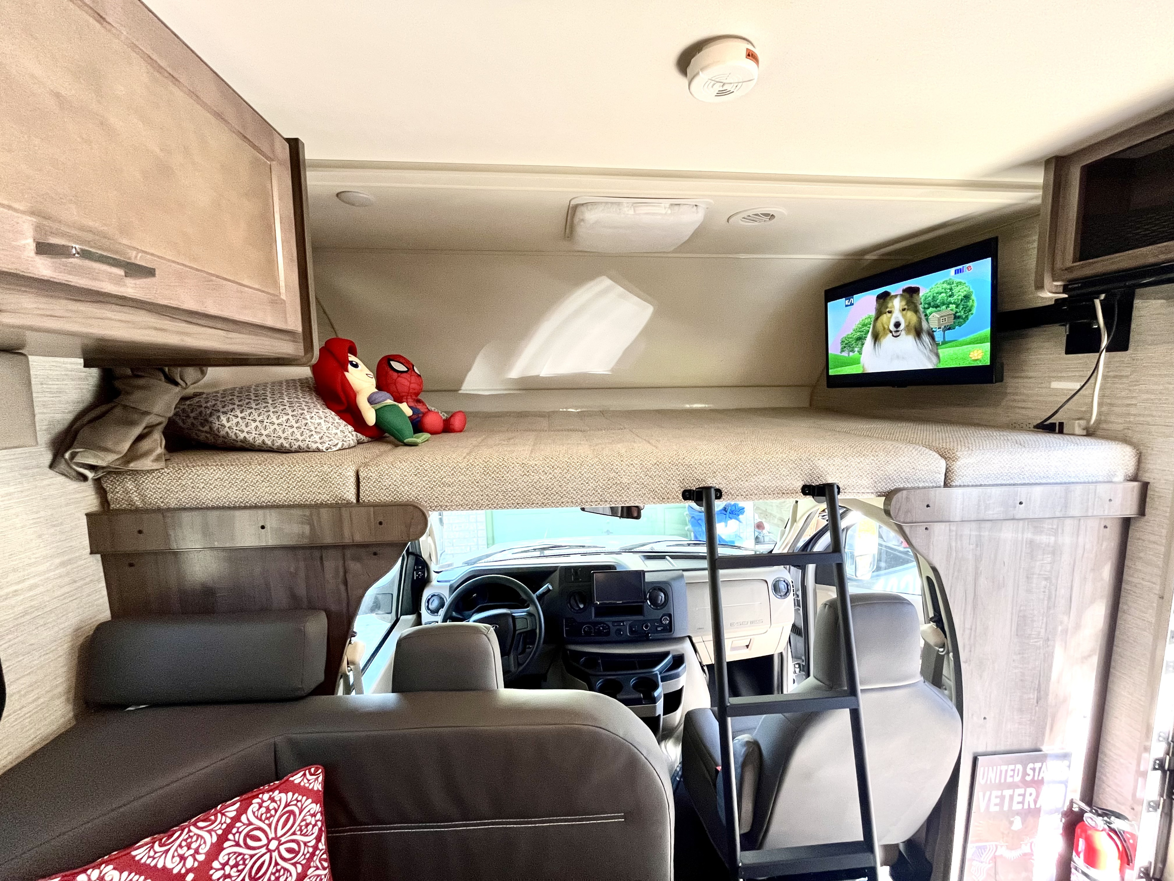 View inside an RV rental showing a cozy cab-over bunk with Ariel & Spider-Man, a TV, and ladder to the driver's area.. Winnebago Minnie Winnie 2021