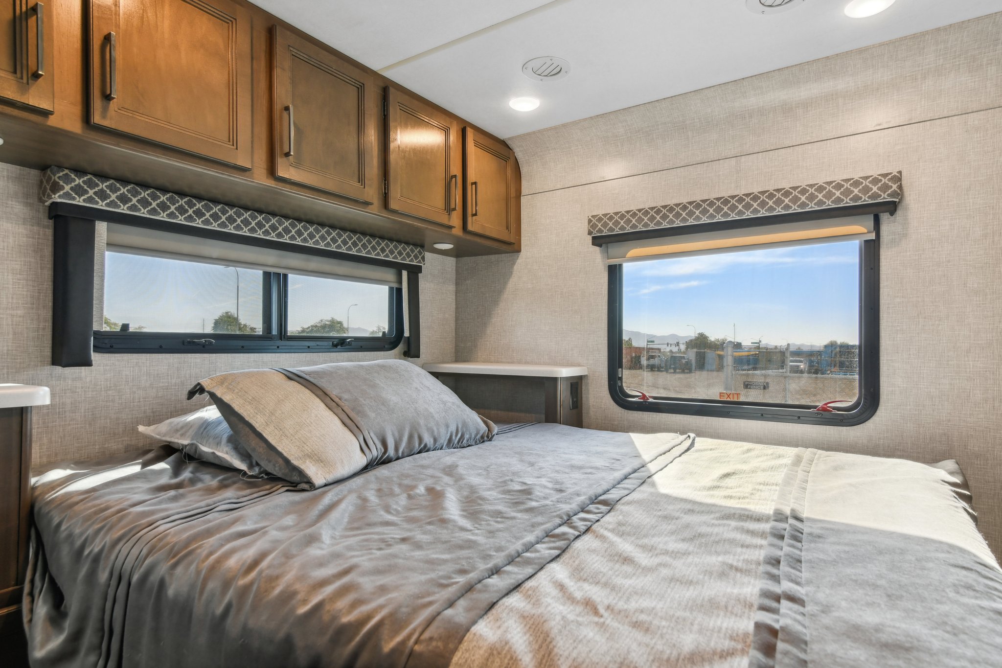 Cozy RV rental bedroom featuring a bed with gray bedding, overhead wooden cabinets, and large windows revealing a sunny outdoor view.. Thor Motor Coach Quantum 2022