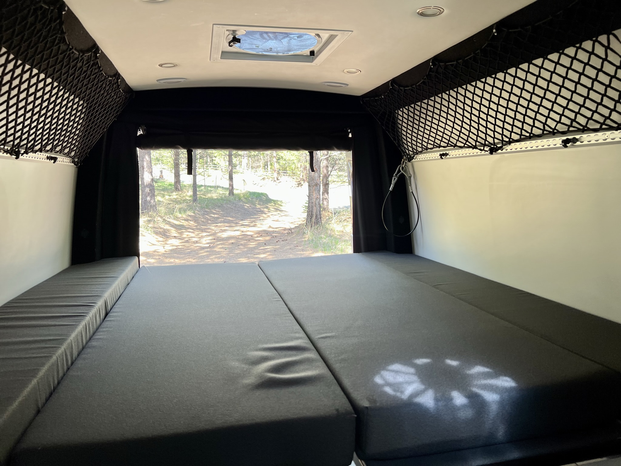 RV rental interior featuring a large, configurable dark gray bed, ceiling fan, storage nets, and a peaceful forest view.. Mercedes-Benz Sprinter RV Motorhome Campervan 2022