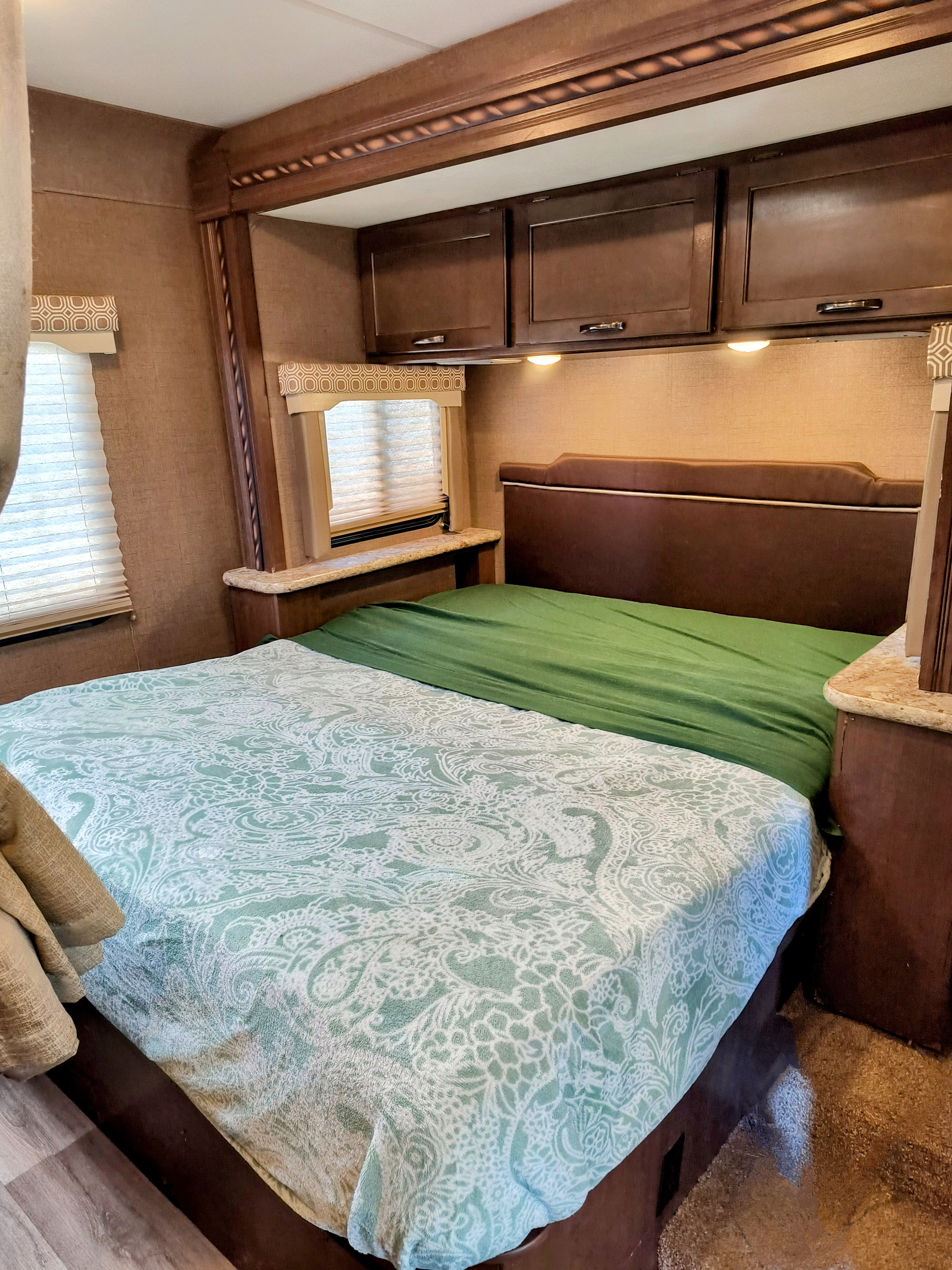 RV rental bedroom: A cozy, compact space with a patterned green bed, dark wood cabinets, and window blinds.. Thor Motor Coach Four Winds 2017