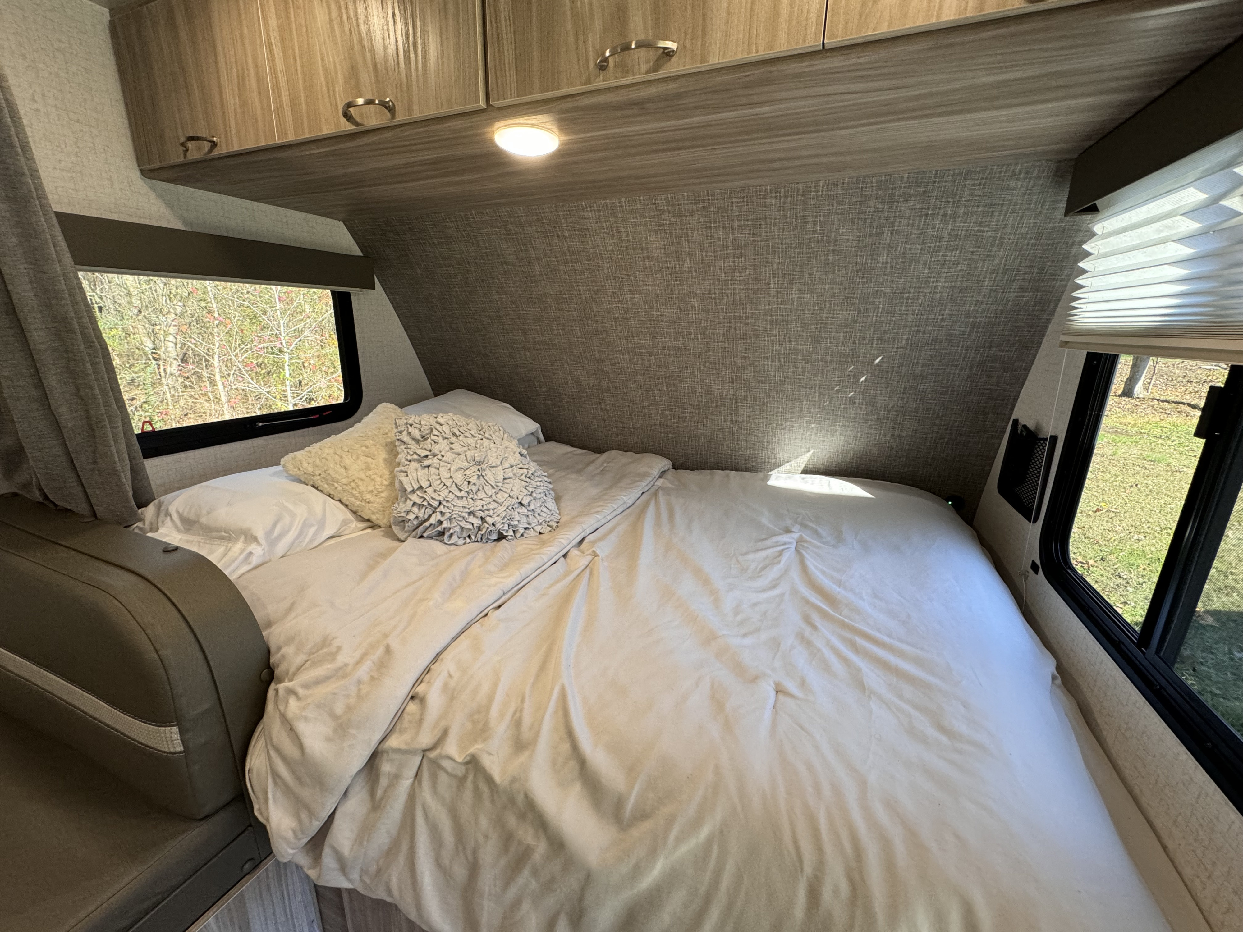 Cozy RV rental bedroom featuring a bed with white linen, overhead cabinets, and windows showing an outdoor forest view.. Winnebago Minnie Winnie 2022