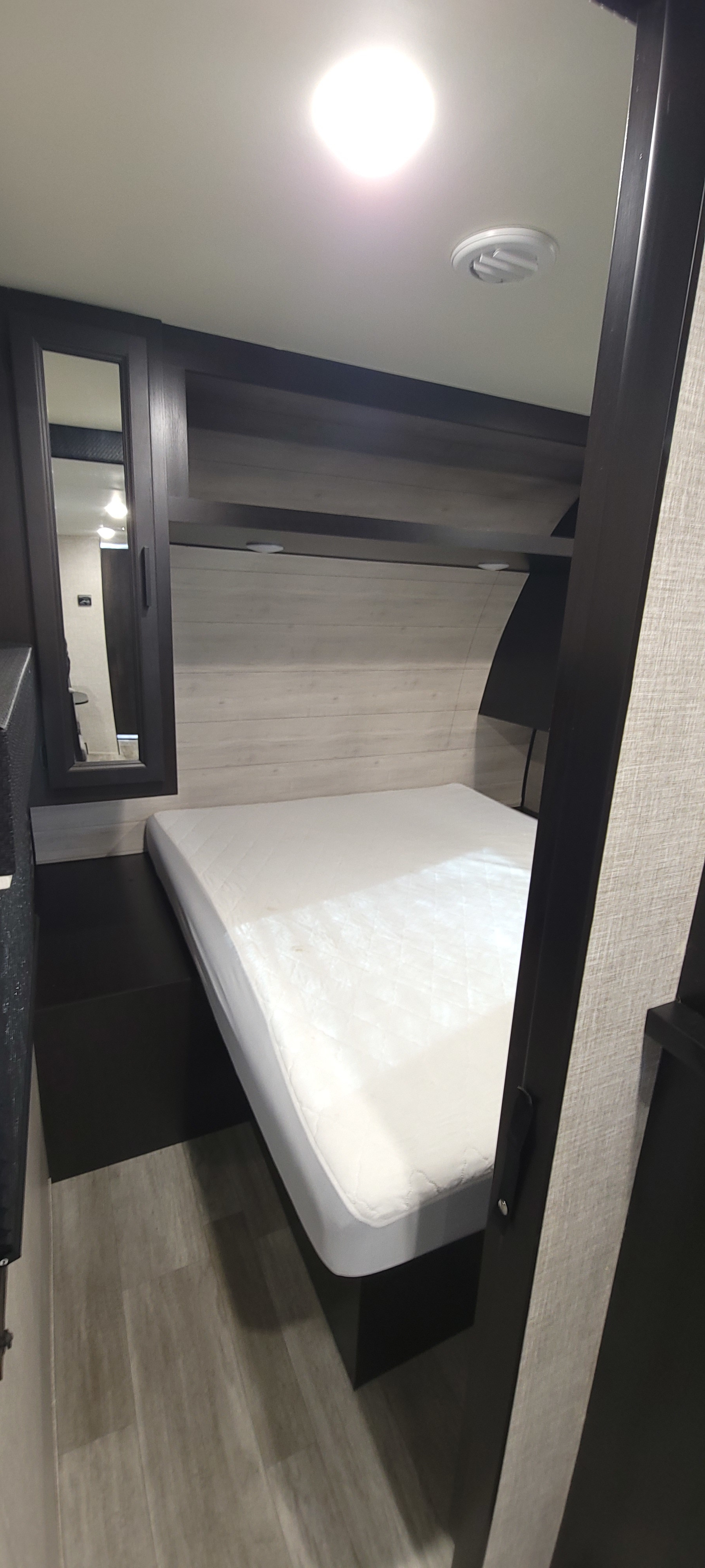 Compact RV rental bedroom featuring a white mattress, dark wood-look cabinetry, light paneled walls, and overhead storage. A mirror is seen on the left.. Jayco Jay Flight Baja 2023