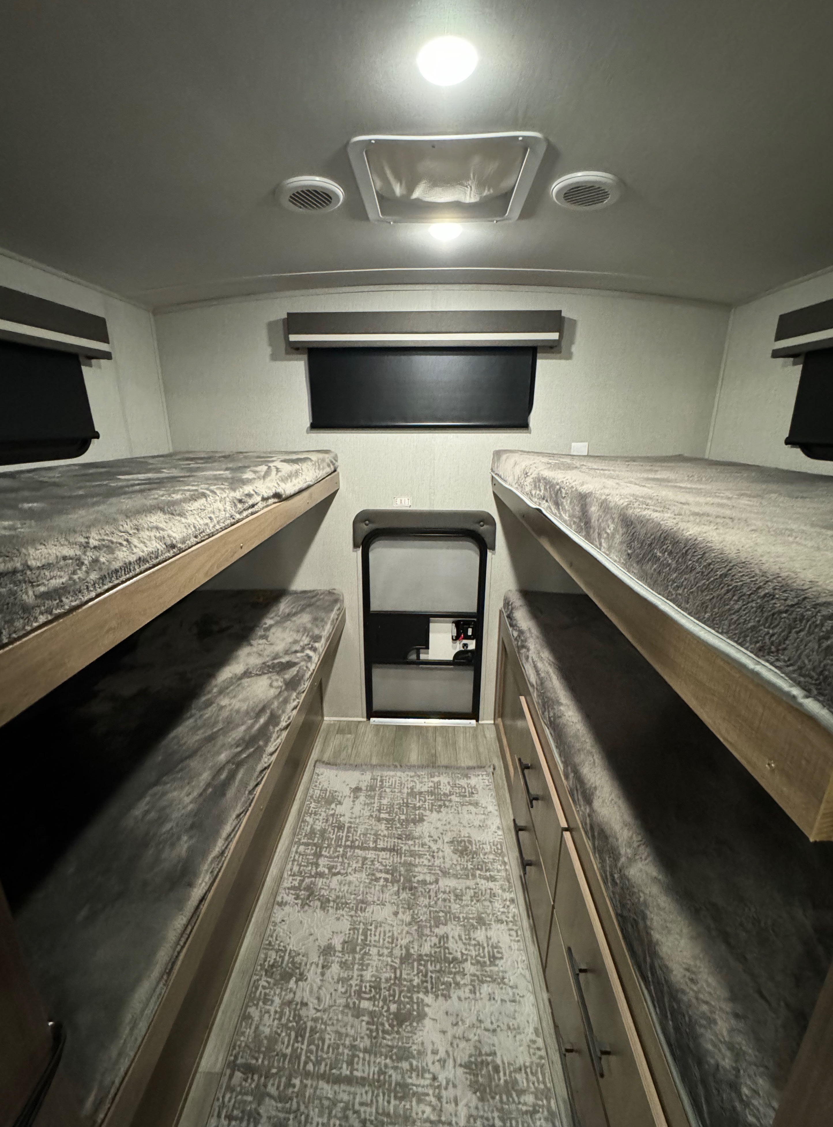 RV rental bunkhouse with four soft beds (two per side), patterned rug, and exit door, illuminated by ceiling lights.. Grand Design Imagine 2023