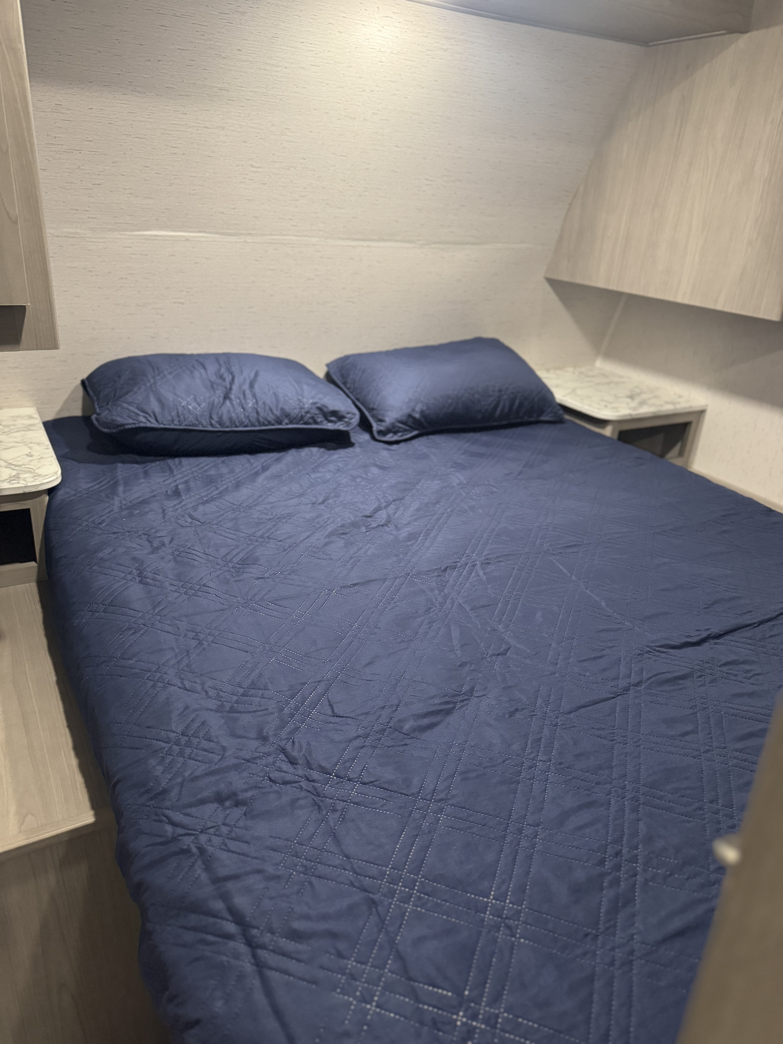 An RV rental bedroom features a navy blue quilted bed with two pillows. Light wood walls and built-in nightstands frame the bed.. Colman Lantern lt 2024