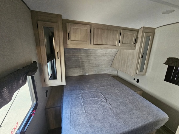An RV rental bedroom featuring a bed with grey linen, light wood cabinets above and beside, and a window for natural light.. Forest River Coachmen Catalina Legacy 2024