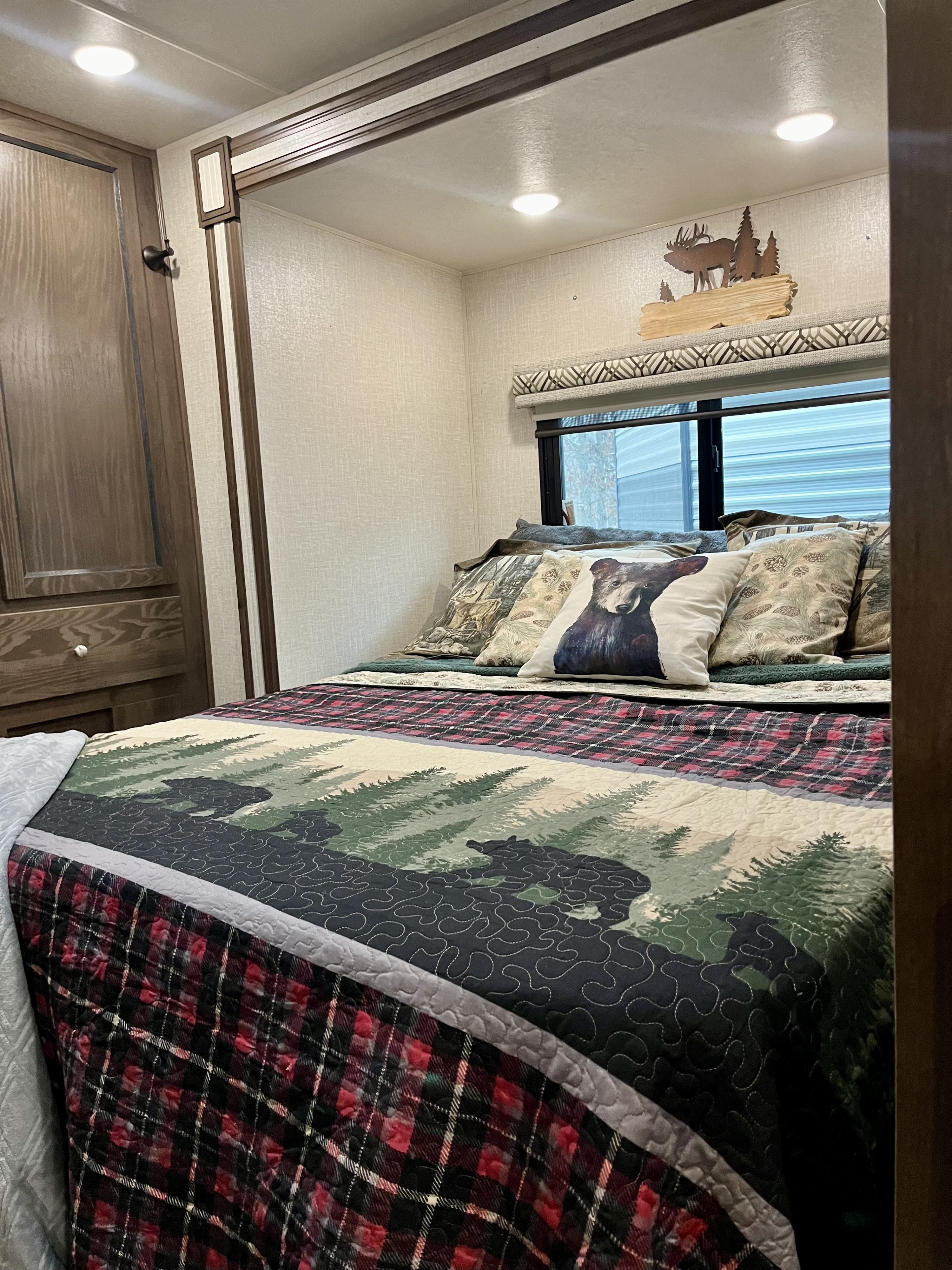 Cozy RV rental bedroom with a rustic bear-themed quilt, window, and wood decor, perfect for a cozy camper stay.. Coachmen Prism 2019