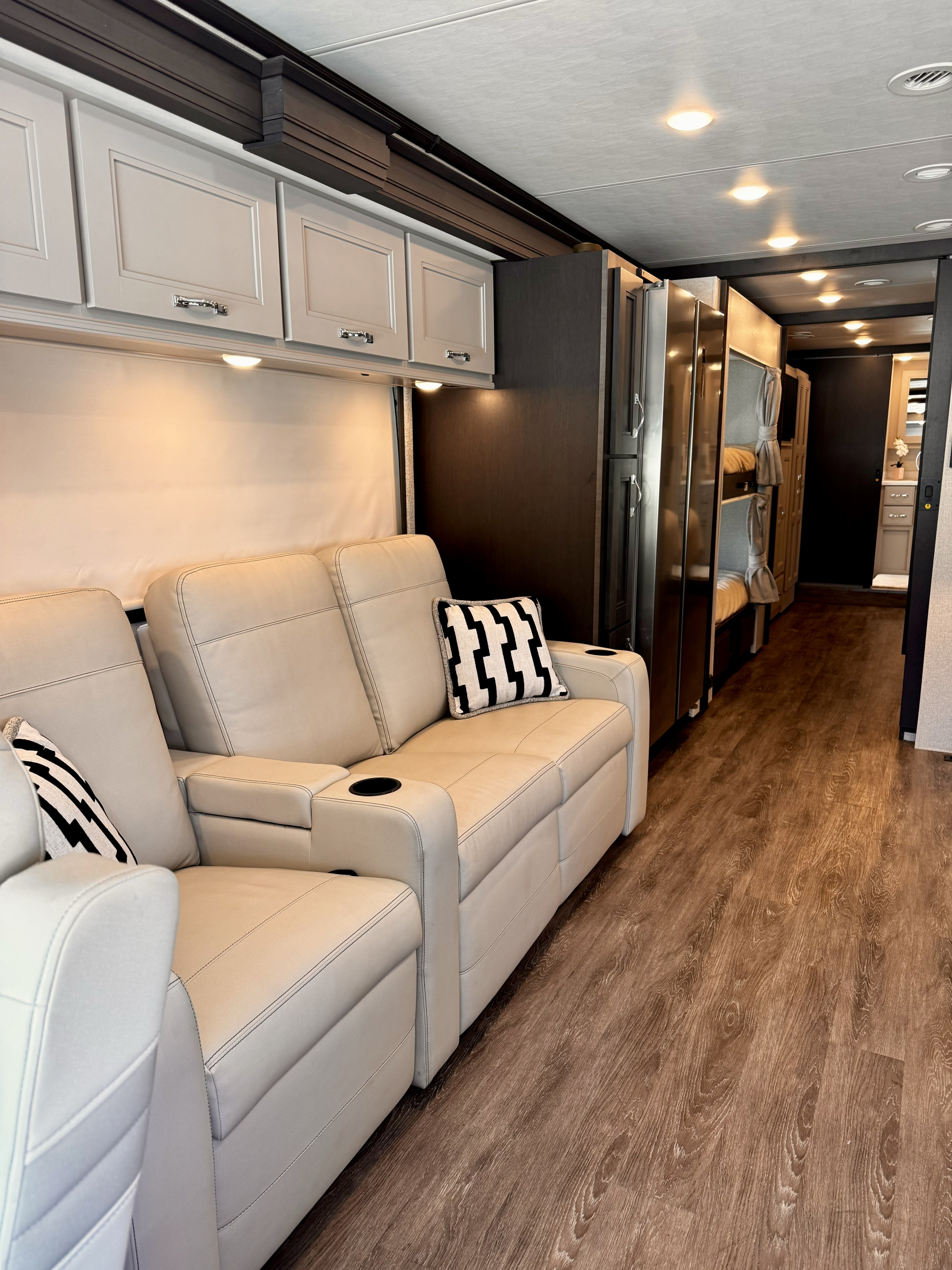 Modern RV rental interior: cream leather recliners with cupholders, overhead cabinets, wood floors, and rear bunk beds.. Thor Riviera 2024
