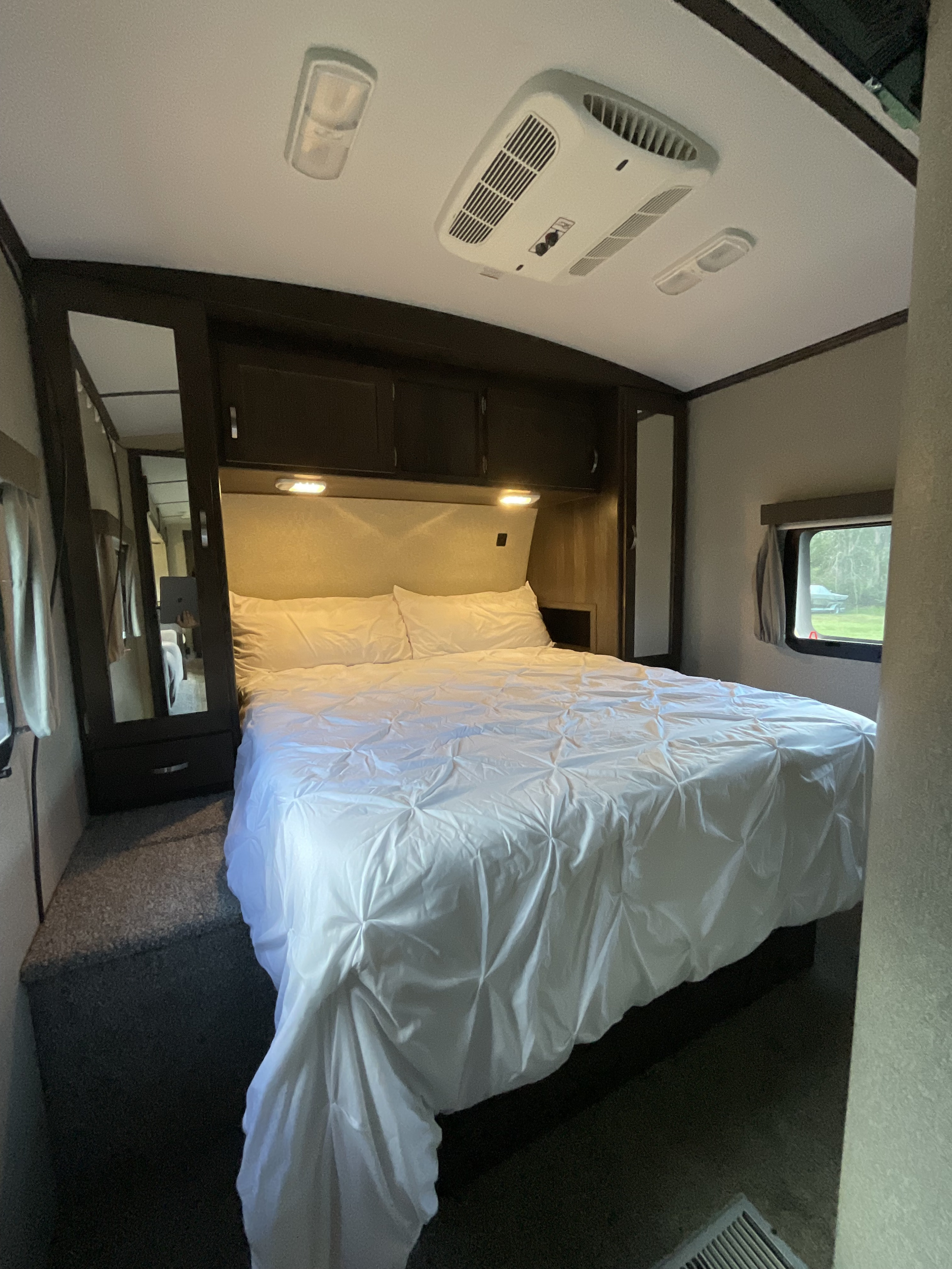 RV rental bedroom features a neatly made bed with white bedding, overhead AC, cabinets, and a window with an outdoor view.. Keystone RV Outback Super-Lite 2018