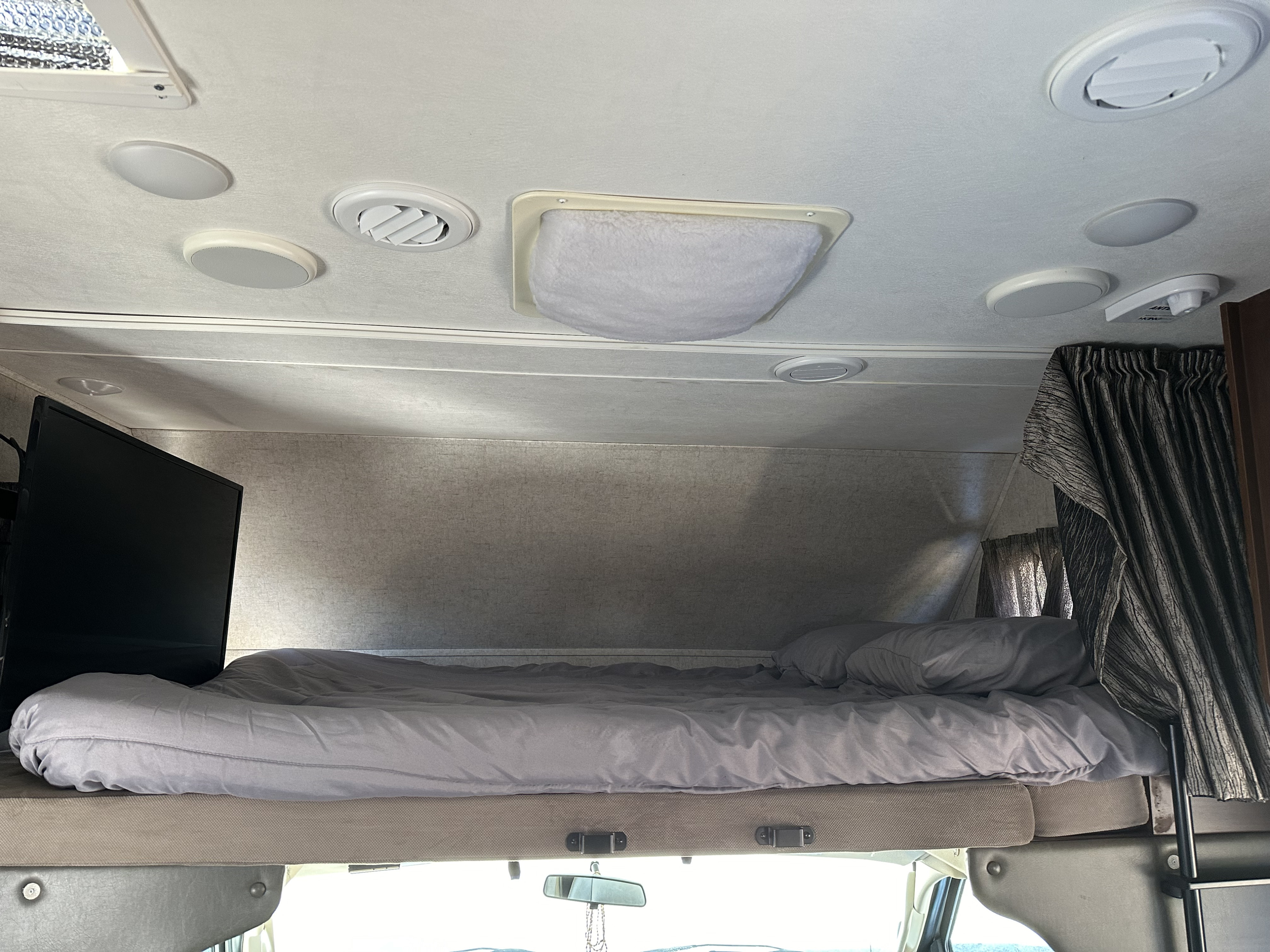 Cozy RV rental overhead bunk bed with grey bedding, mounted TV, ceiling lights, and vents. Ideal RV sleeping space.. Forest River Sunseeker 2018