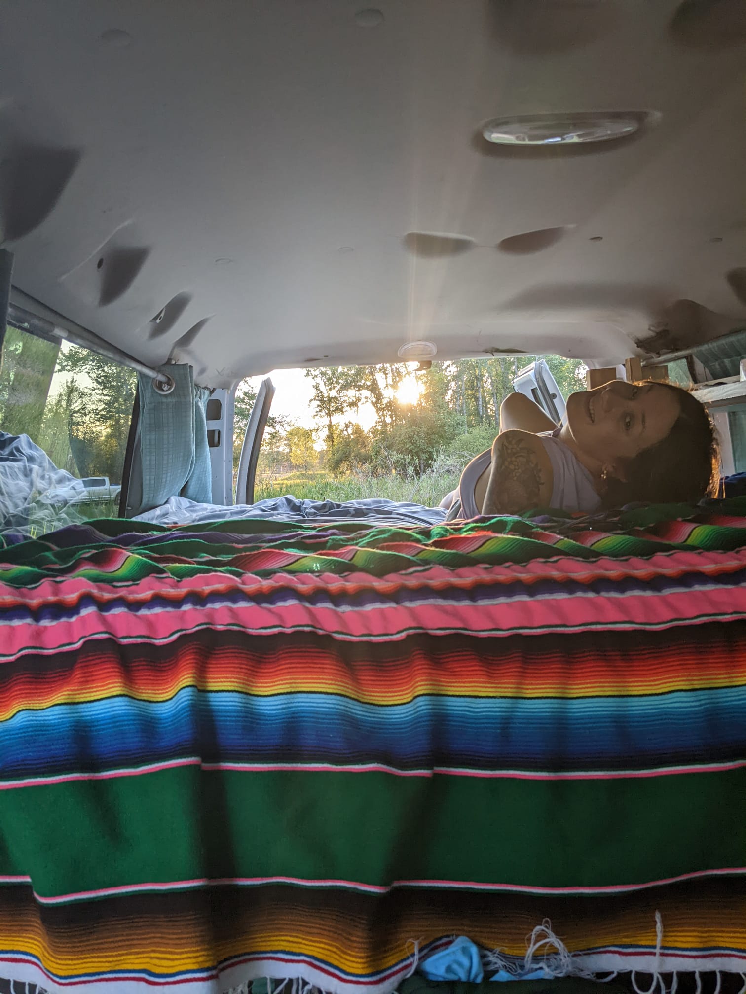 Relaxing in an RV rental with a colorful blanket & sunset view. A woman smiles, enjoying van life in nature.. Ford Glampervan 2014