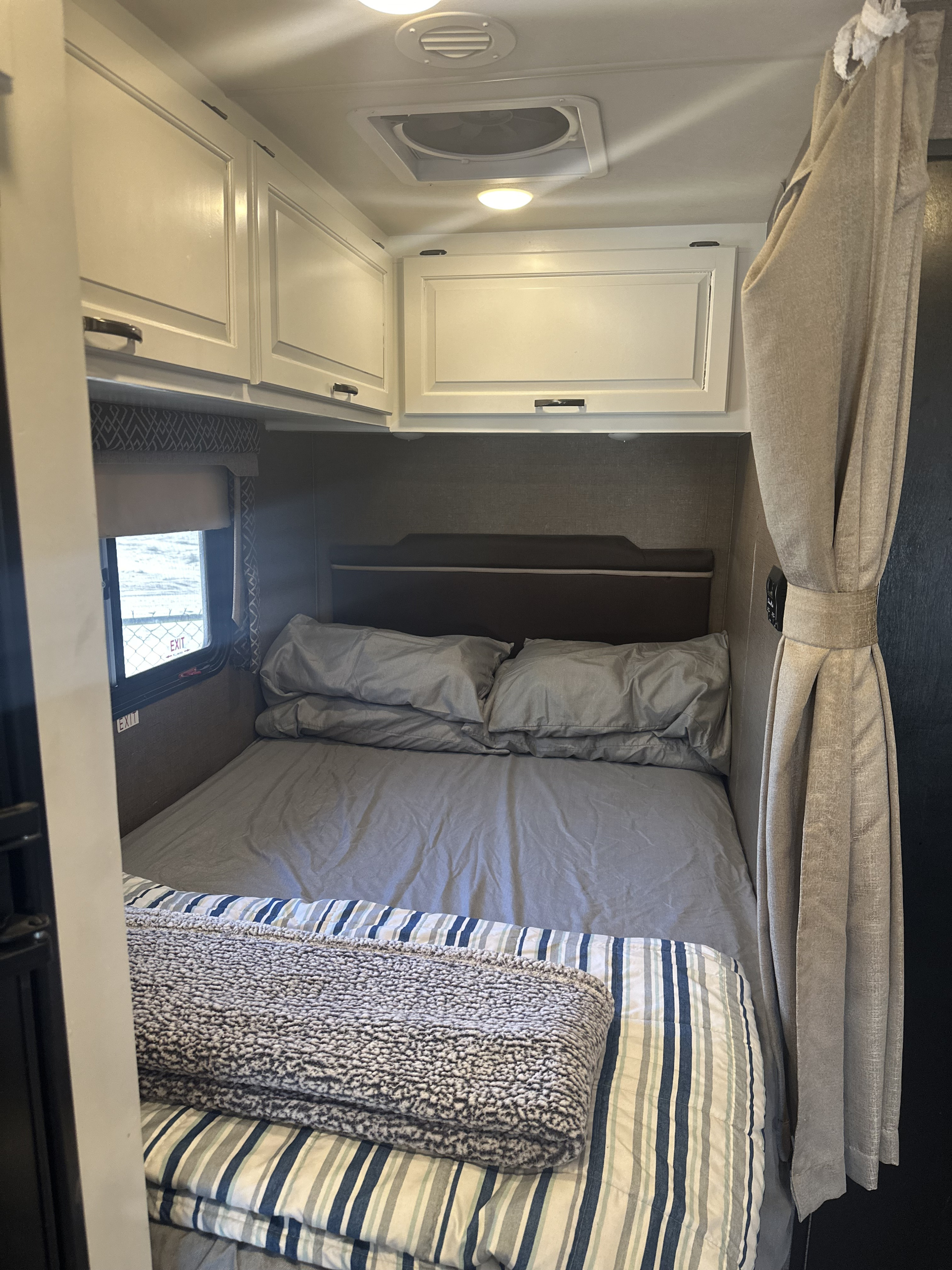 Cozy RV rental bedroom with a bed, gray sheets, striped blanket, overhead cabinets, and an exit window on the left.. Thor Four Winds 2018