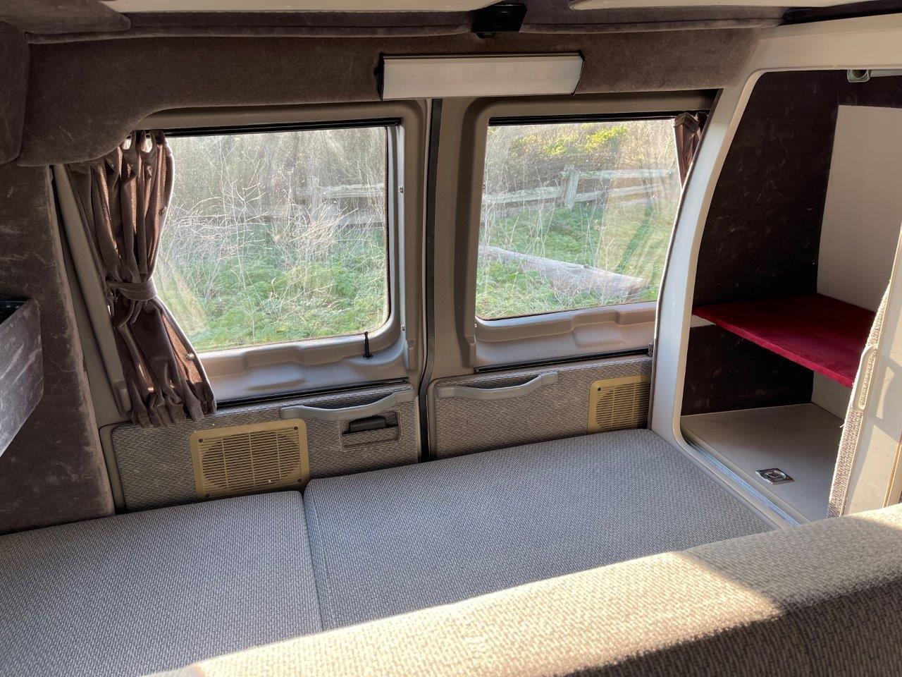 Interior of an RV rental showing a comfy bed, windows with curtains, and built-in storage. Ideal for road trips!. Ford E350 2008