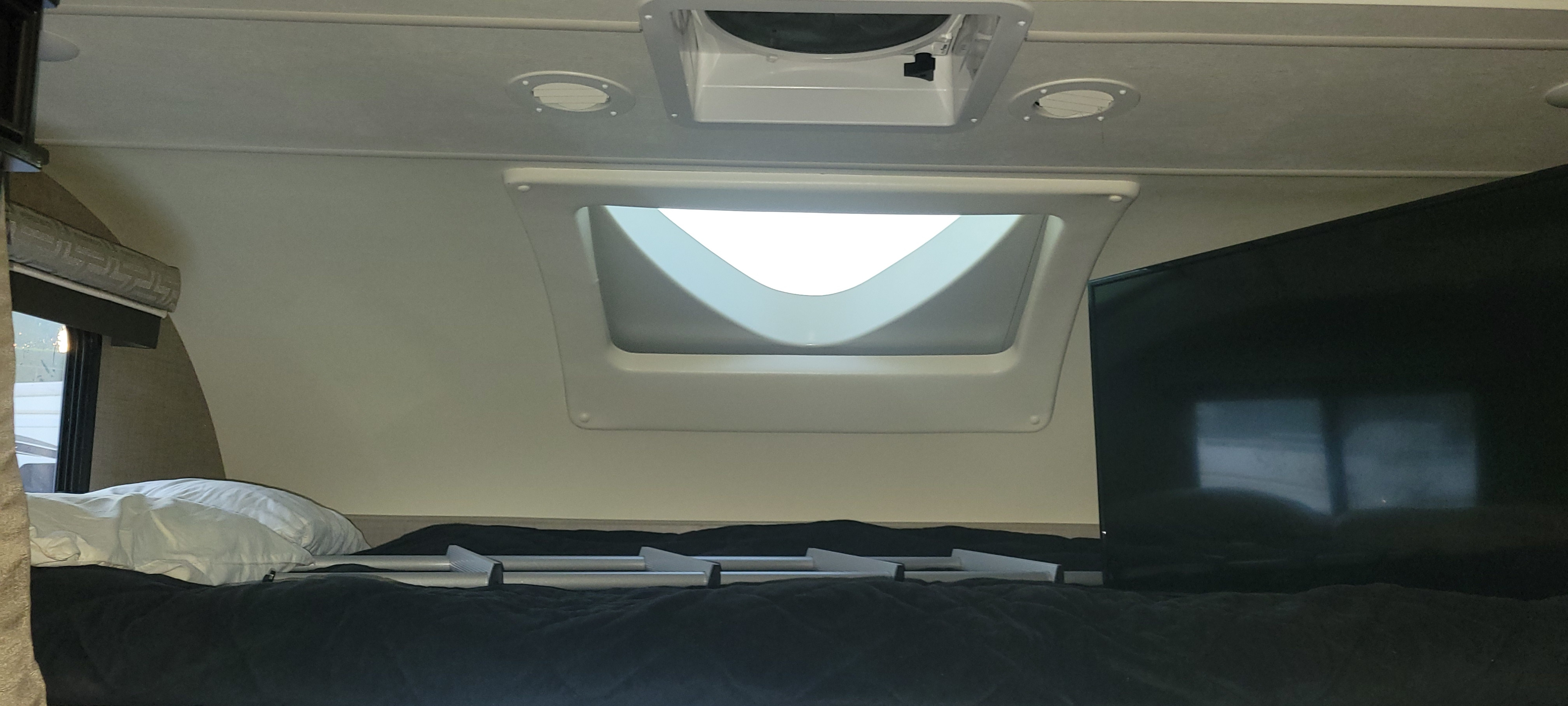 Inside an RV rental: a cozy cab-over bunk bed with bright skylight, ceiling fan, TV, and silver bed supports.. Thor Quantum 2017
