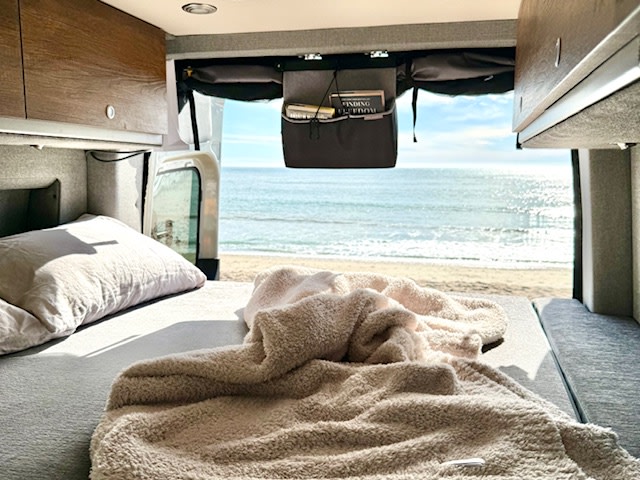 RV rental bedroom: A cozy bed inside a camper van, offering a stunning view of a sunny beach and ocean.. Storyteller Overland Storyteller Overland Class B 2023