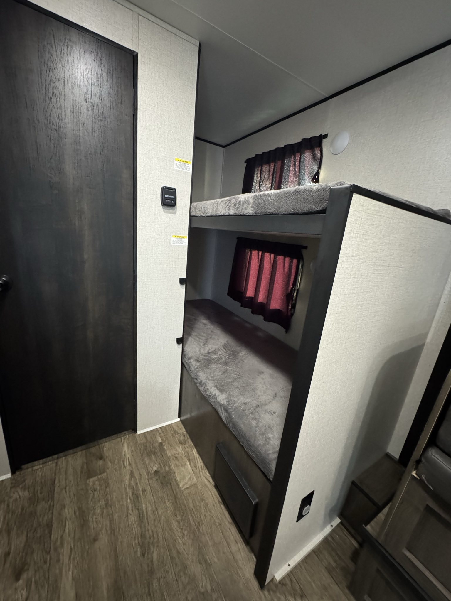 Interior of an RV rental showing cozy bunk beds with grey blankets, red window curtains, and wood flooring.. Starcraft Autumn Ridge 2022