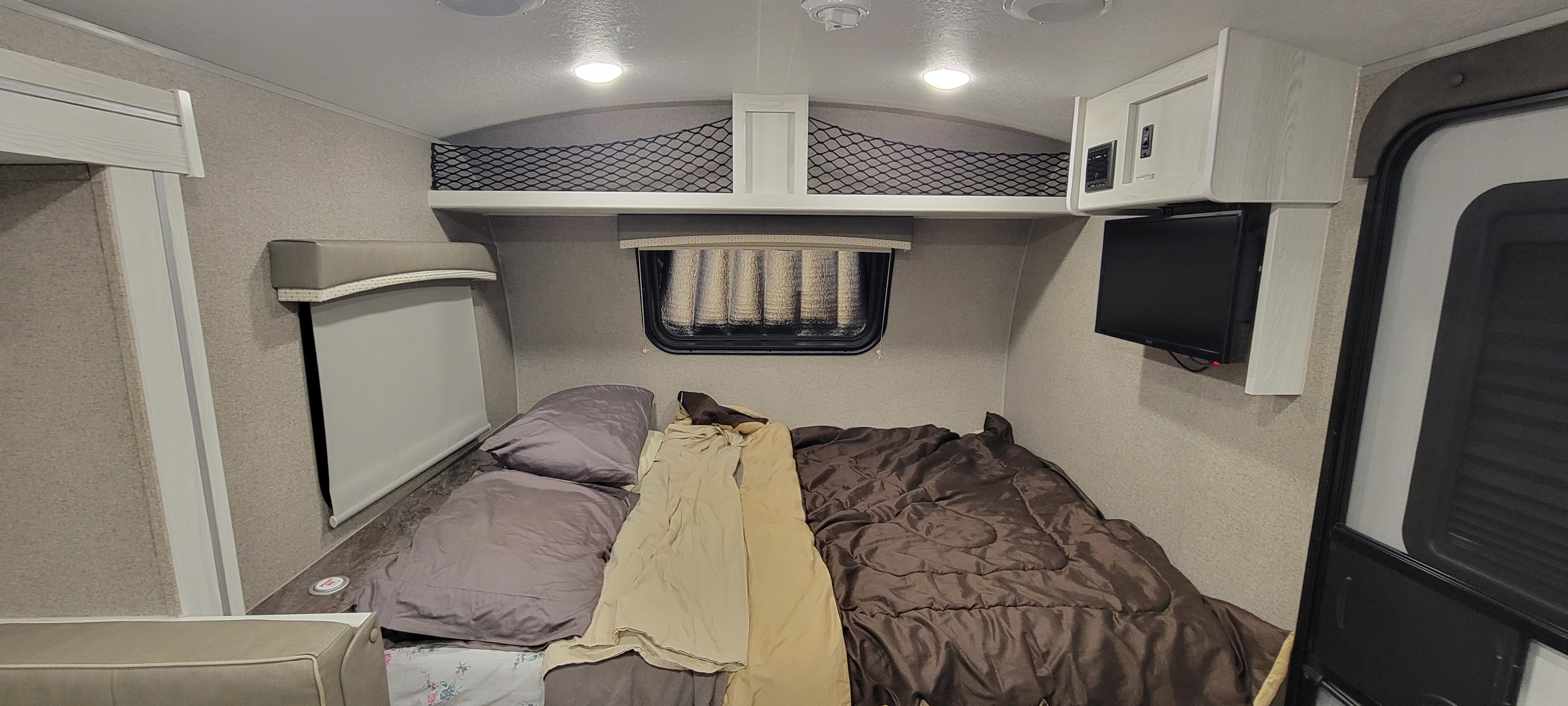 Inside a compact RV rental, showing a neatly made bed with pillows, an overhead storage net, window, TV, and entrance door.. Forest River Rockwood Geo Pro 2020