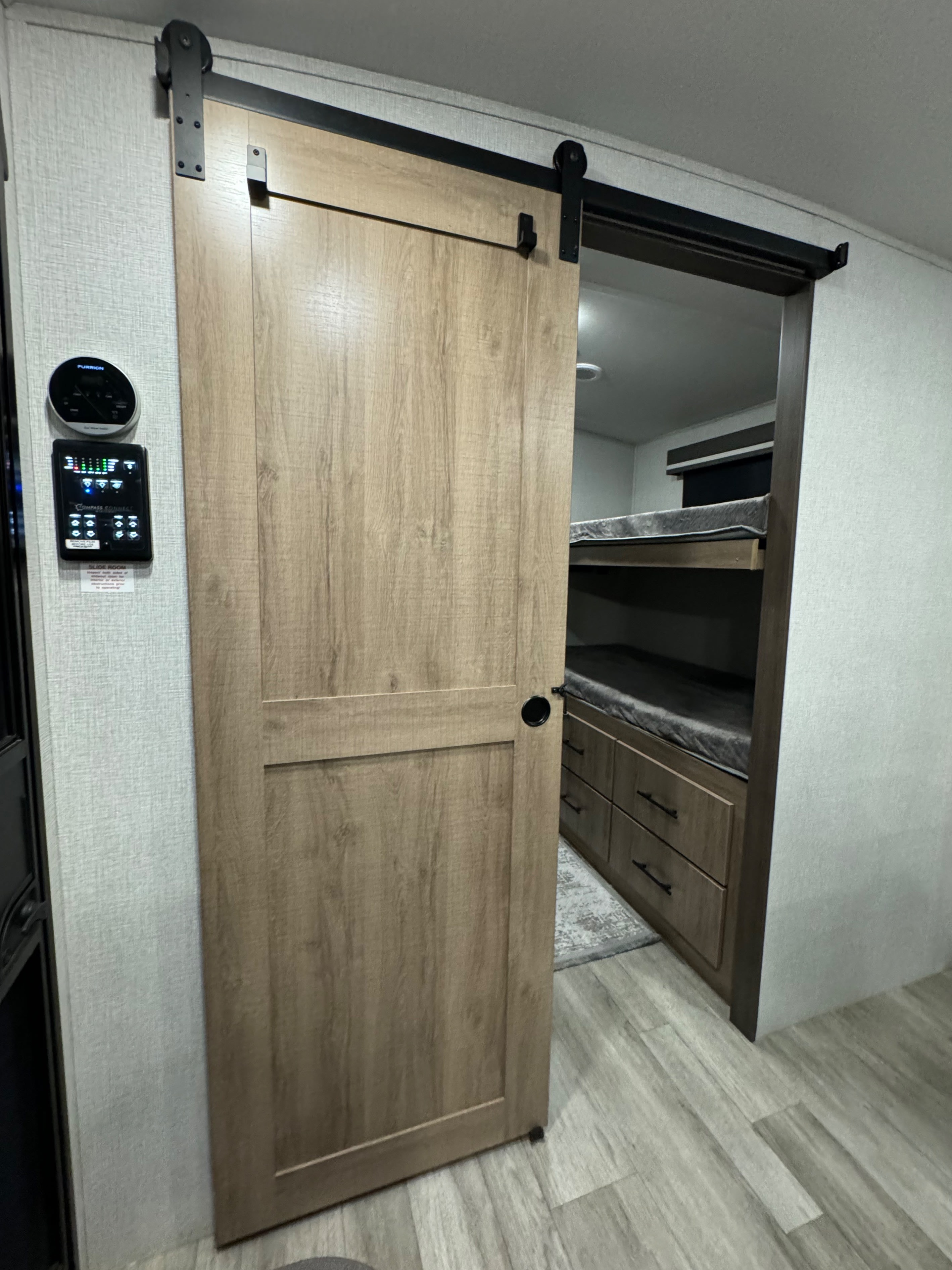 Modern RV rental interior with a wood-grain sliding barn door revealing a bunk room with built-in drawers. Wall-mounted controls visible.. Grand Design Imagine 2023