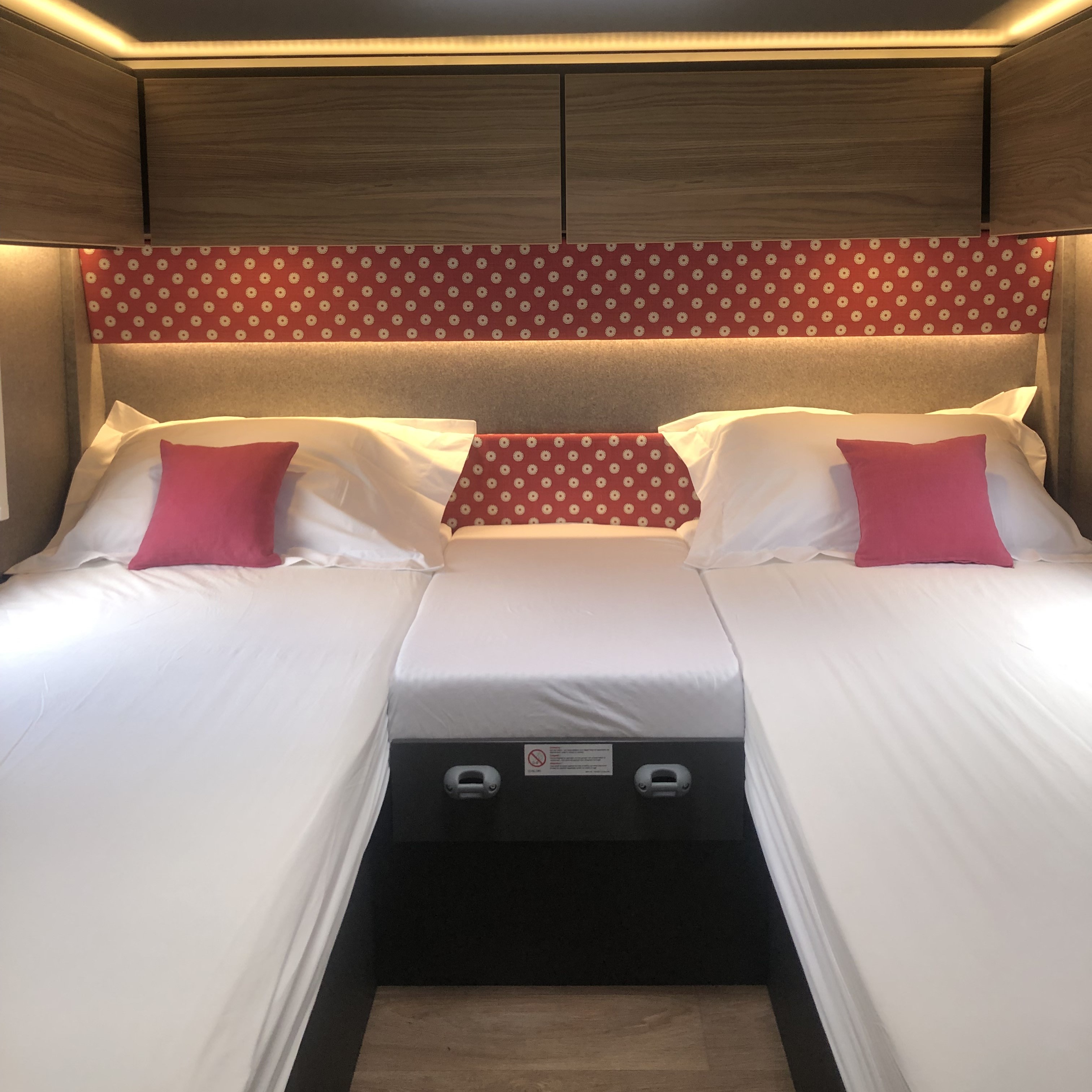 Bright and cozy sleeping area in an RV rental, showing twin beds, overhead storage, and soft headboard lighting.. GlamperRV DBL 2026