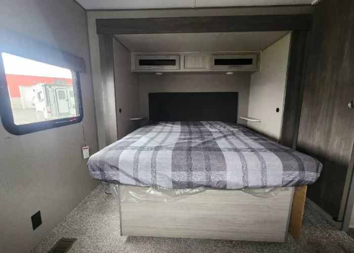 Spacious RV rental bedroom with a comfy plaid bed, overhead cabinets, and window view. Perfect for your next adventure!. Forest River Catalina 2022