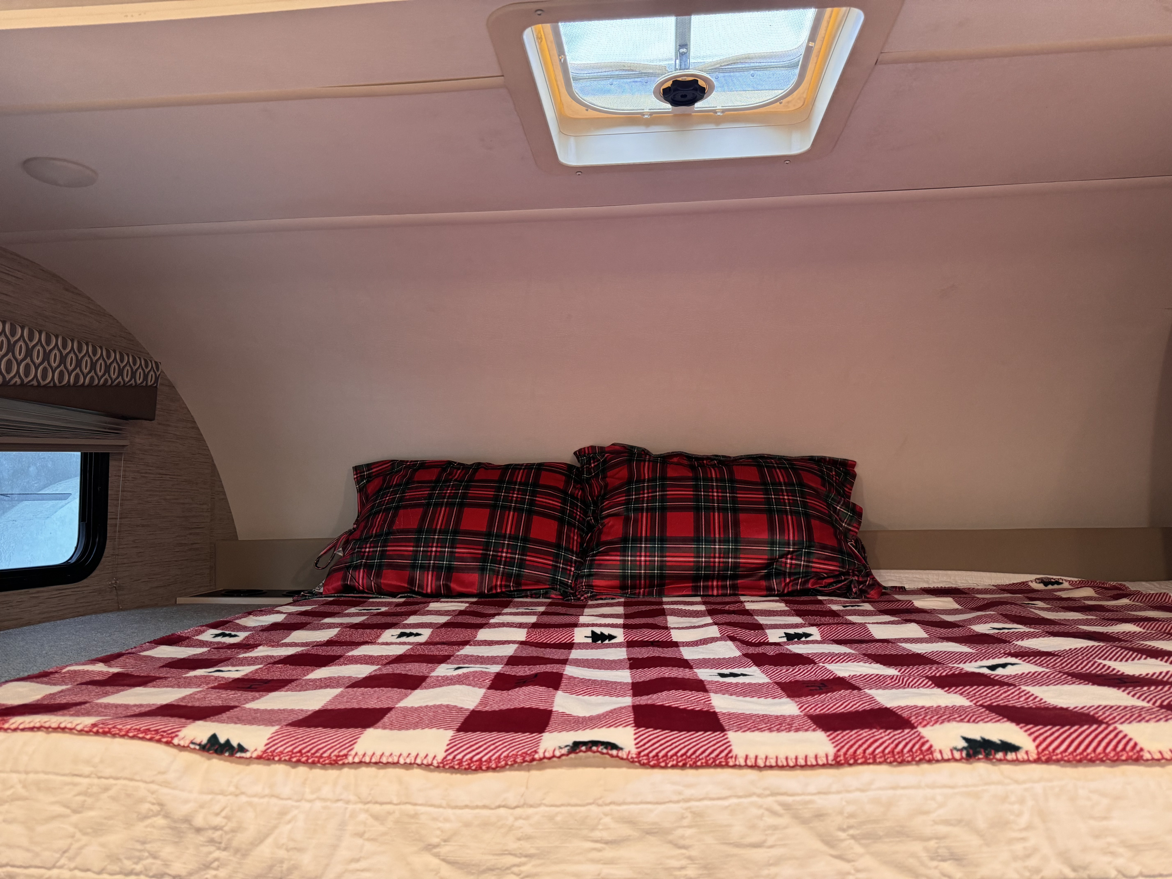 Inside a cozy RV rental: a comfortable bed with festive plaid pillows & a tree-patterned blanket. Window shows snow.. Thor Freedom Elite 2022