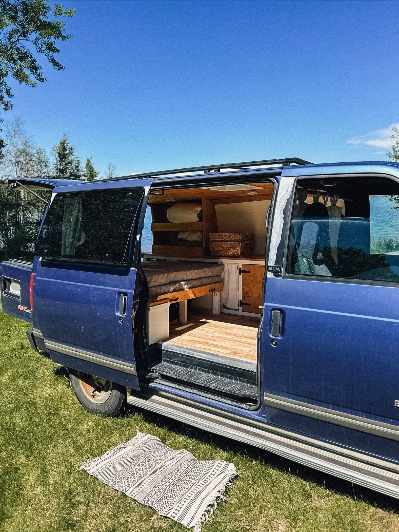 This blue conversion van, perfect for an RV rental, shows its custom wood-finished interior with a bed and storage. Parked on grass.. Gmc Safari 1996