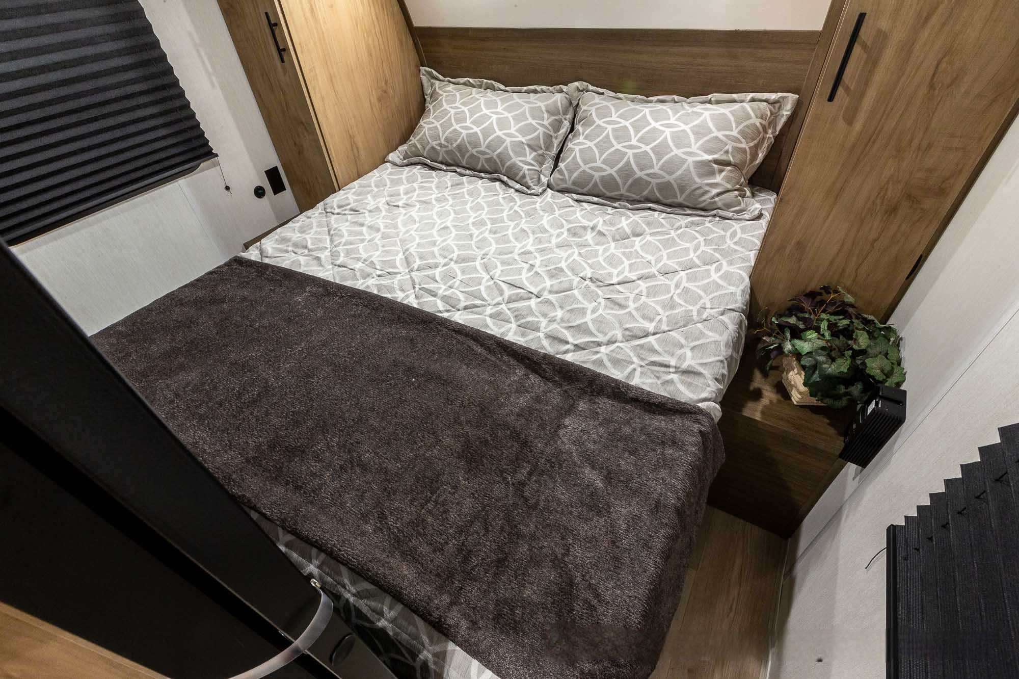 Compact RV rental bedroom: patterned bed, pillows, wooden storage, and window blind for cozy travel.. Forest River 23dbh 2025