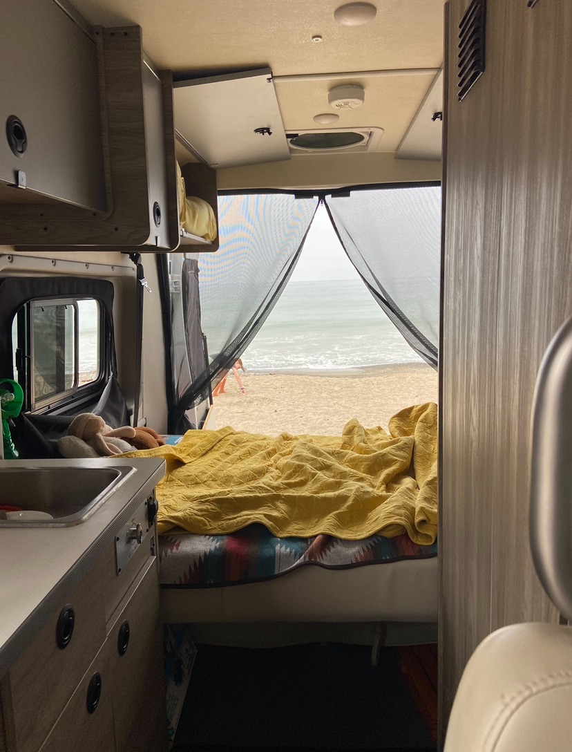 Inside an RV rental, a comfy bed and kitchenette face a stunning beach and ocean view, seen through a mesh screen.. Dodge Winnebago 2023