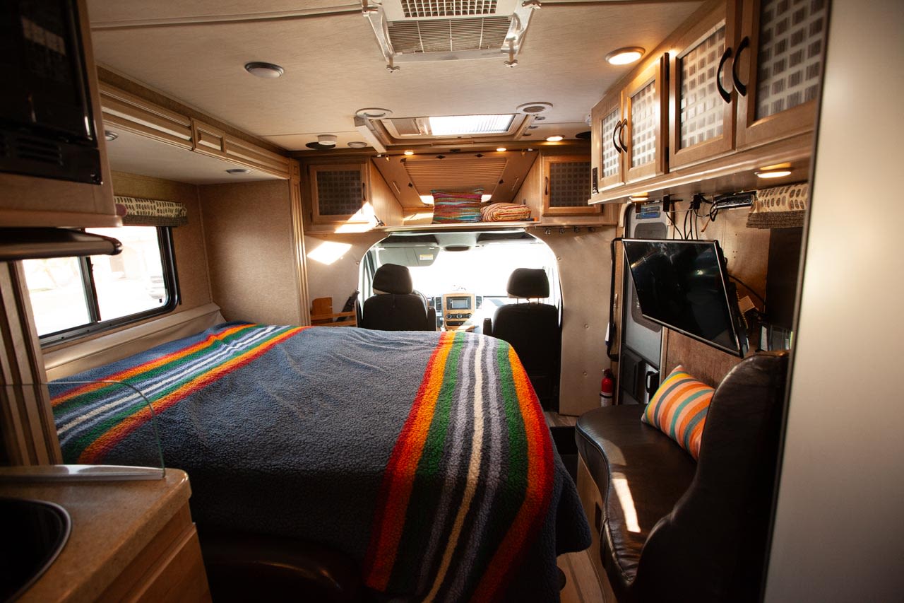 Bright RV rental interior showing a bed with a vibrant blanket, kitchen, TV, and front cabin area.. Mercedes Prism 2015