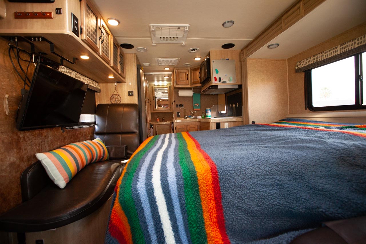 Cozy RV rental interior showing bedroom with colorful blanket, living area, and kitchen setup.. Mercedes Prism 2015