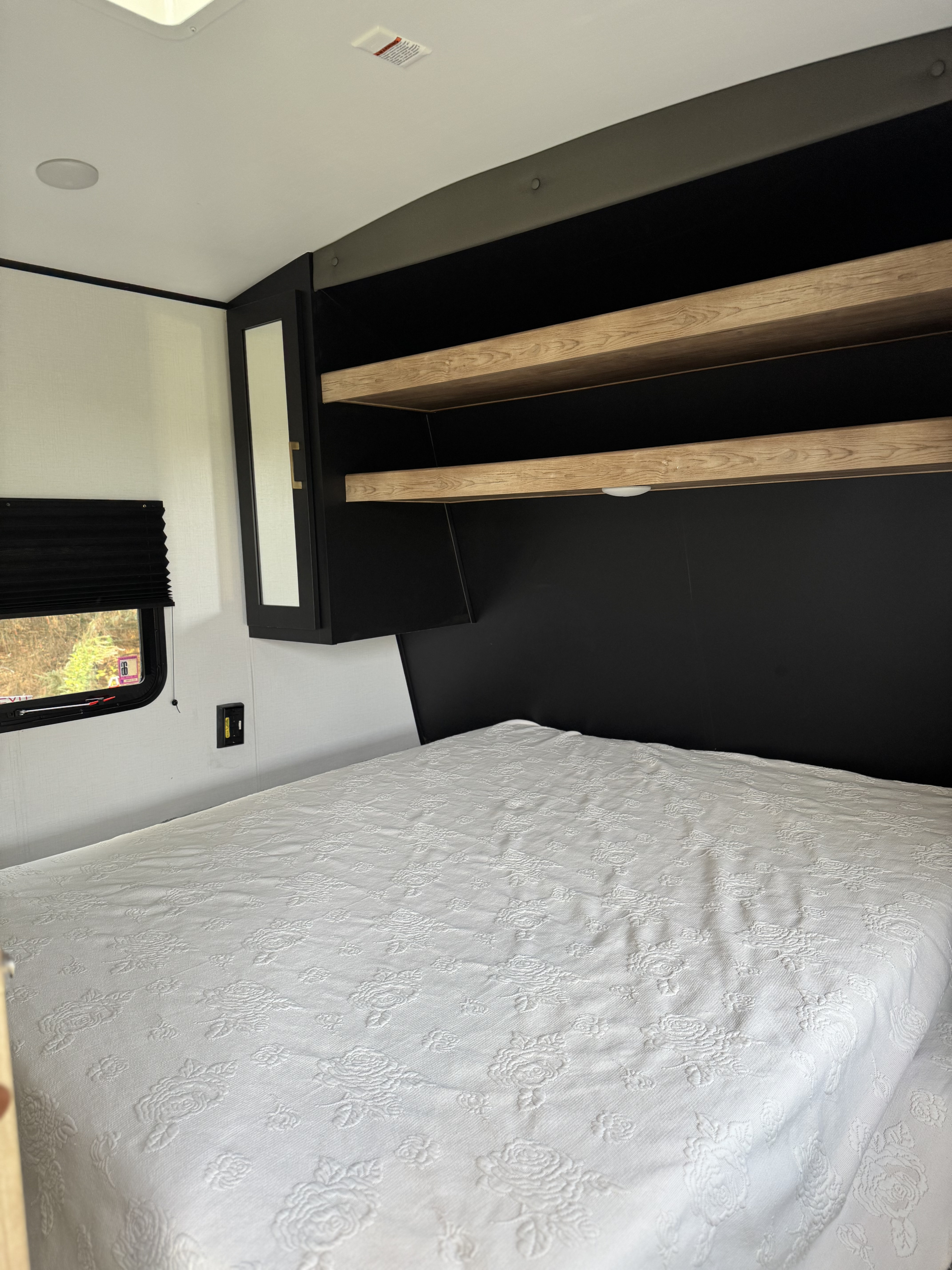 RV rental bedroom featuring a bed with a white embossed floral bedspread, black storage, and window view.. Forest River Springdale 2022