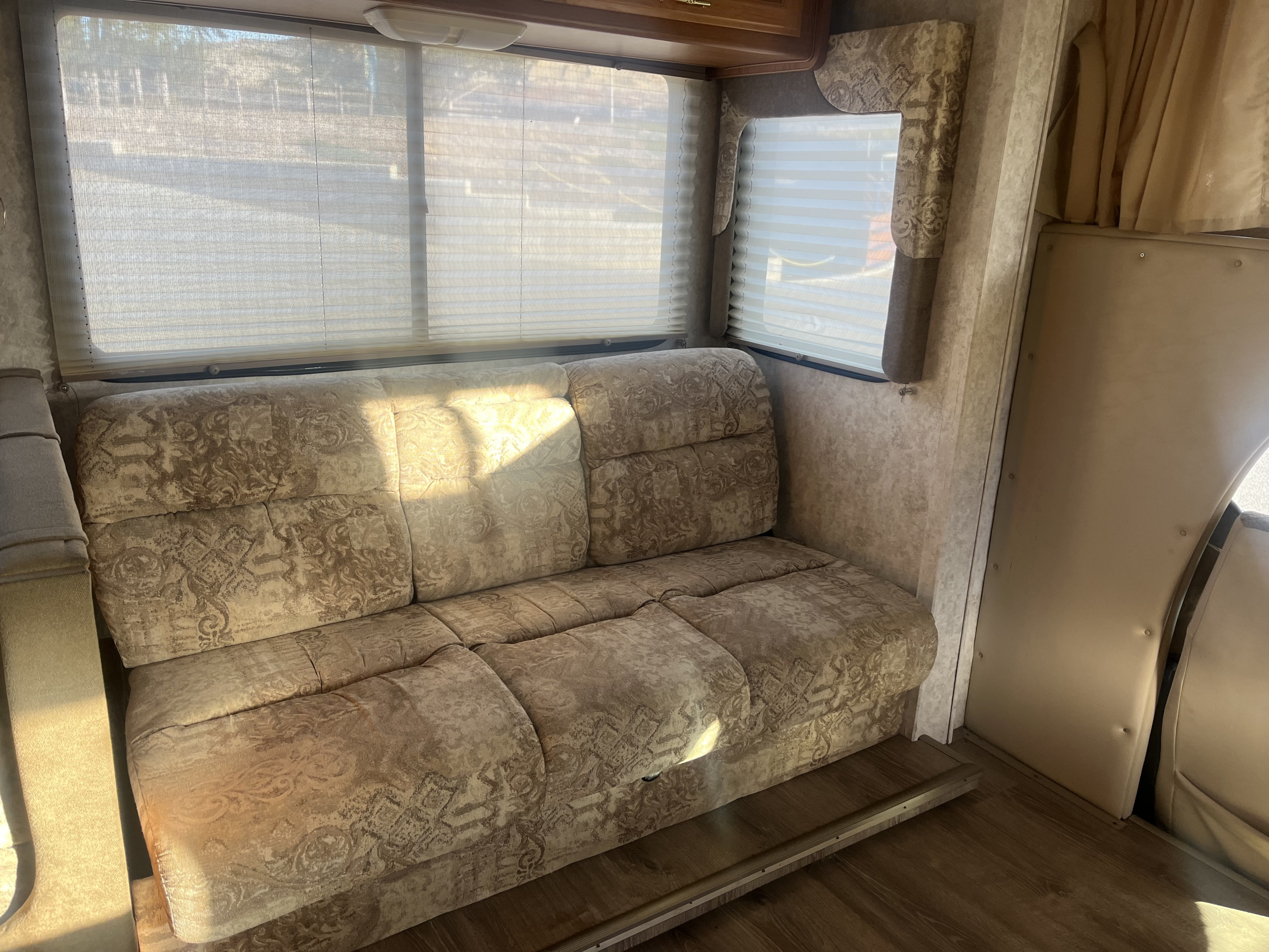 Cozy RV rental interior featuring a comfortable patterned sofa, light-filtering window blinds, and wood flooring bathed in sunlight.. Tioga Tioga Motorhome 2000