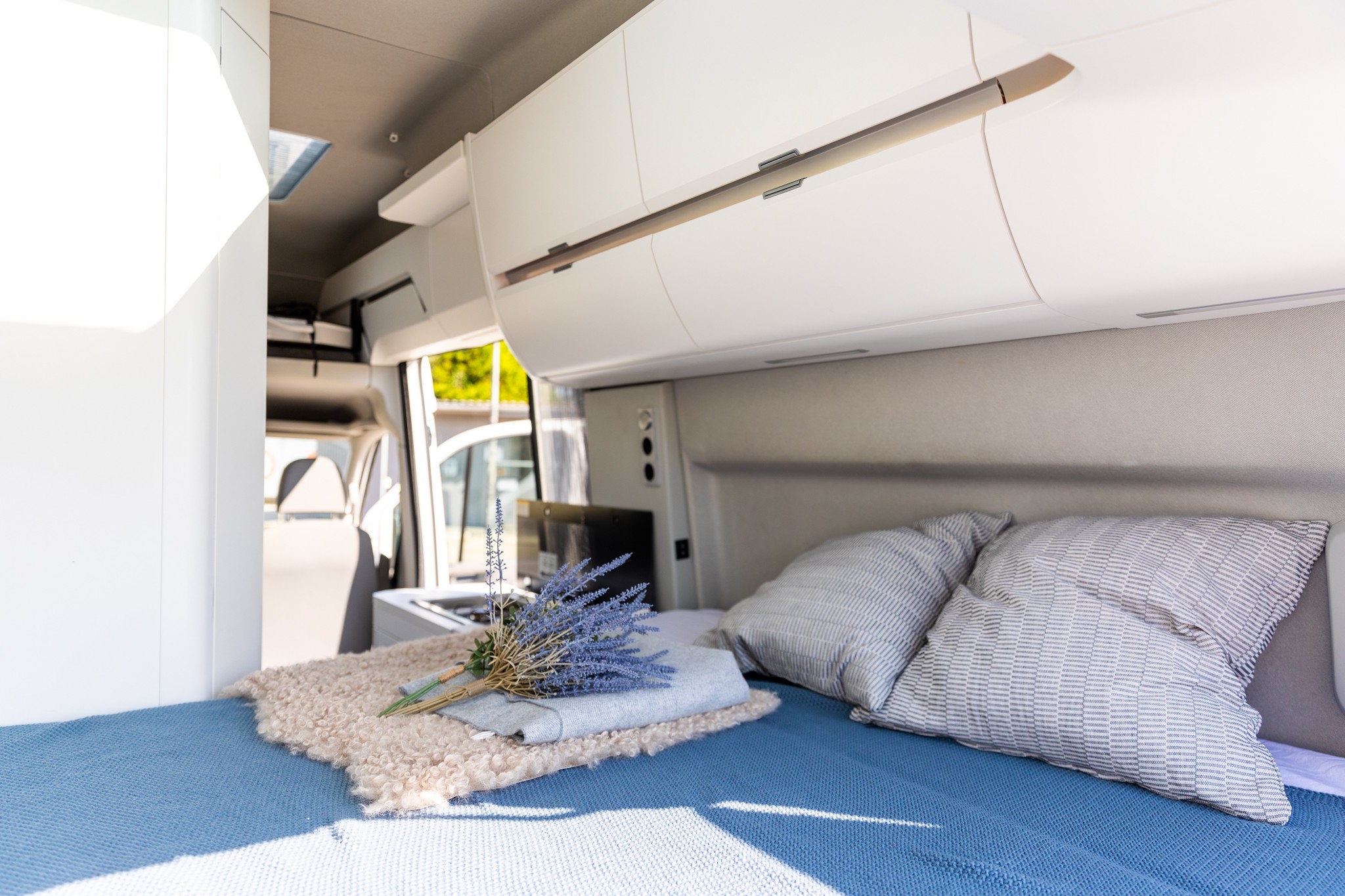 Bright RV rental interior showing a neatly made bed with blue bedding, striped pillows, and lavender decor. Overhead storage is visible.. Volkswagen Grand California 2022