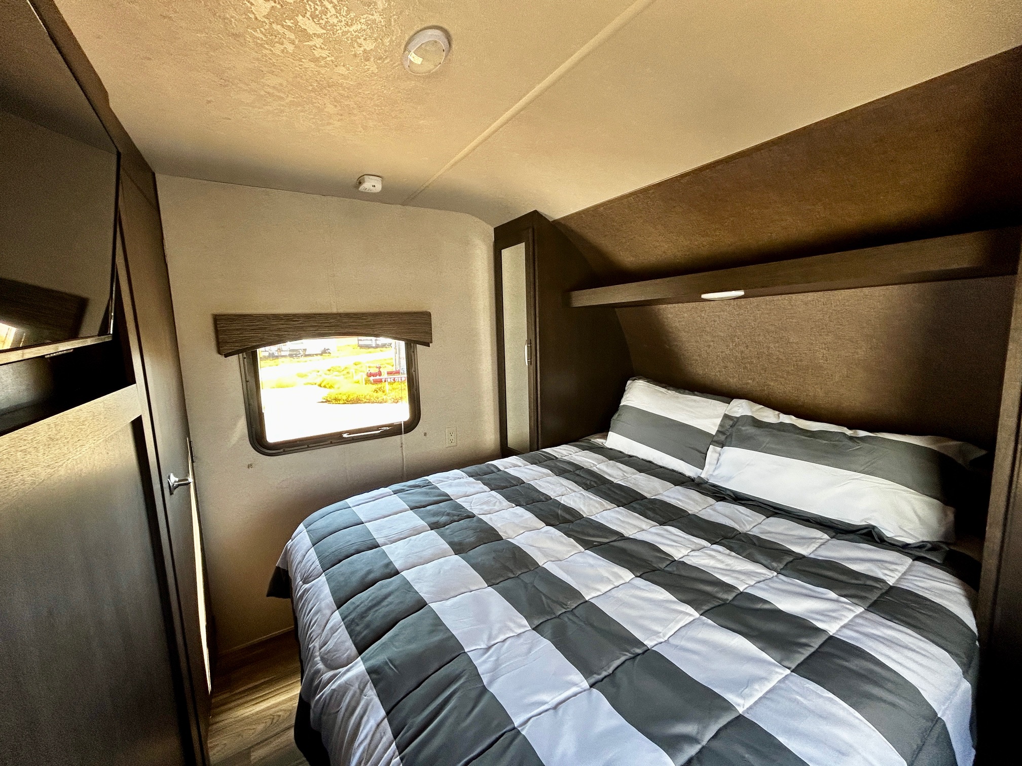 Inside an RV rental: A comfortable bedroom with a gray and white checkered comforter, window view, and dark wood storage.. Forest River Evo 2017