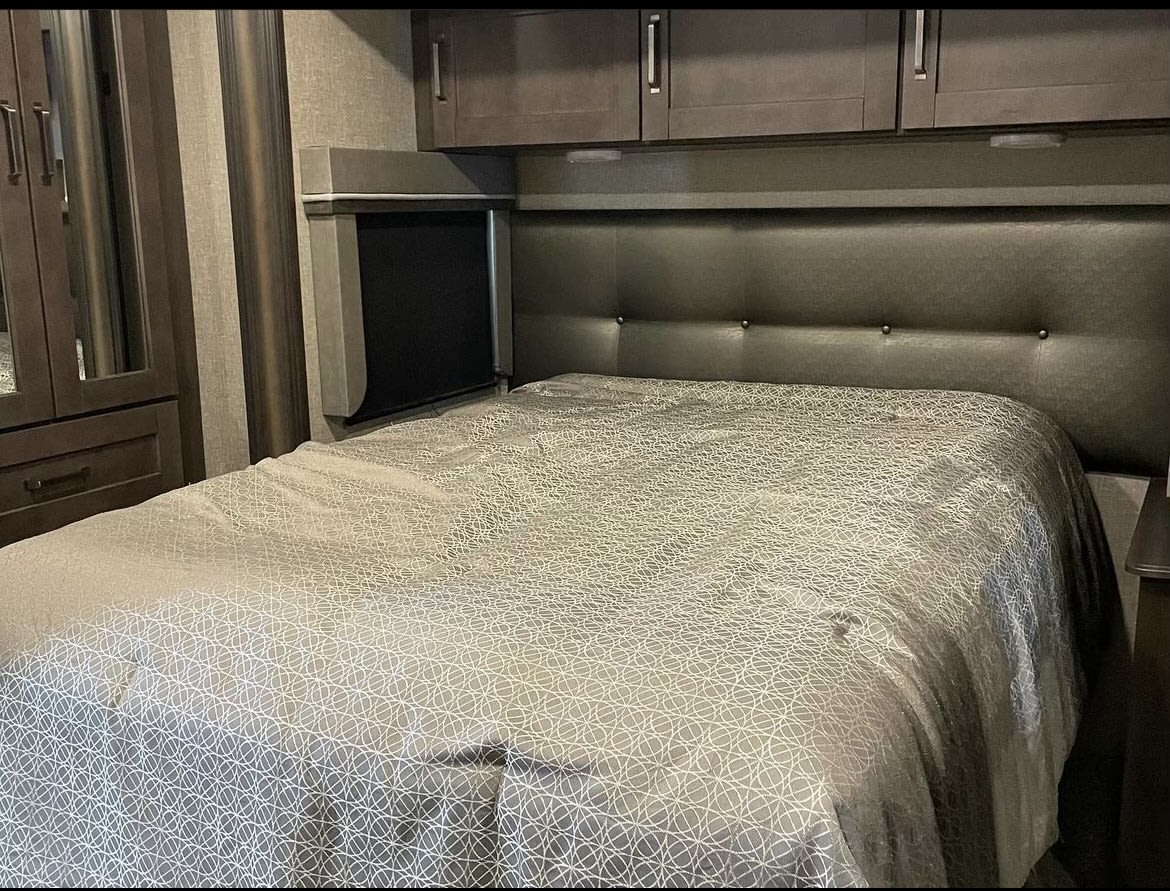 Inside an RV rental: a modern bedroom with a grey patterned bedspread, dark padded headboard, and overhead storage.. Grand Design Reflection 2020