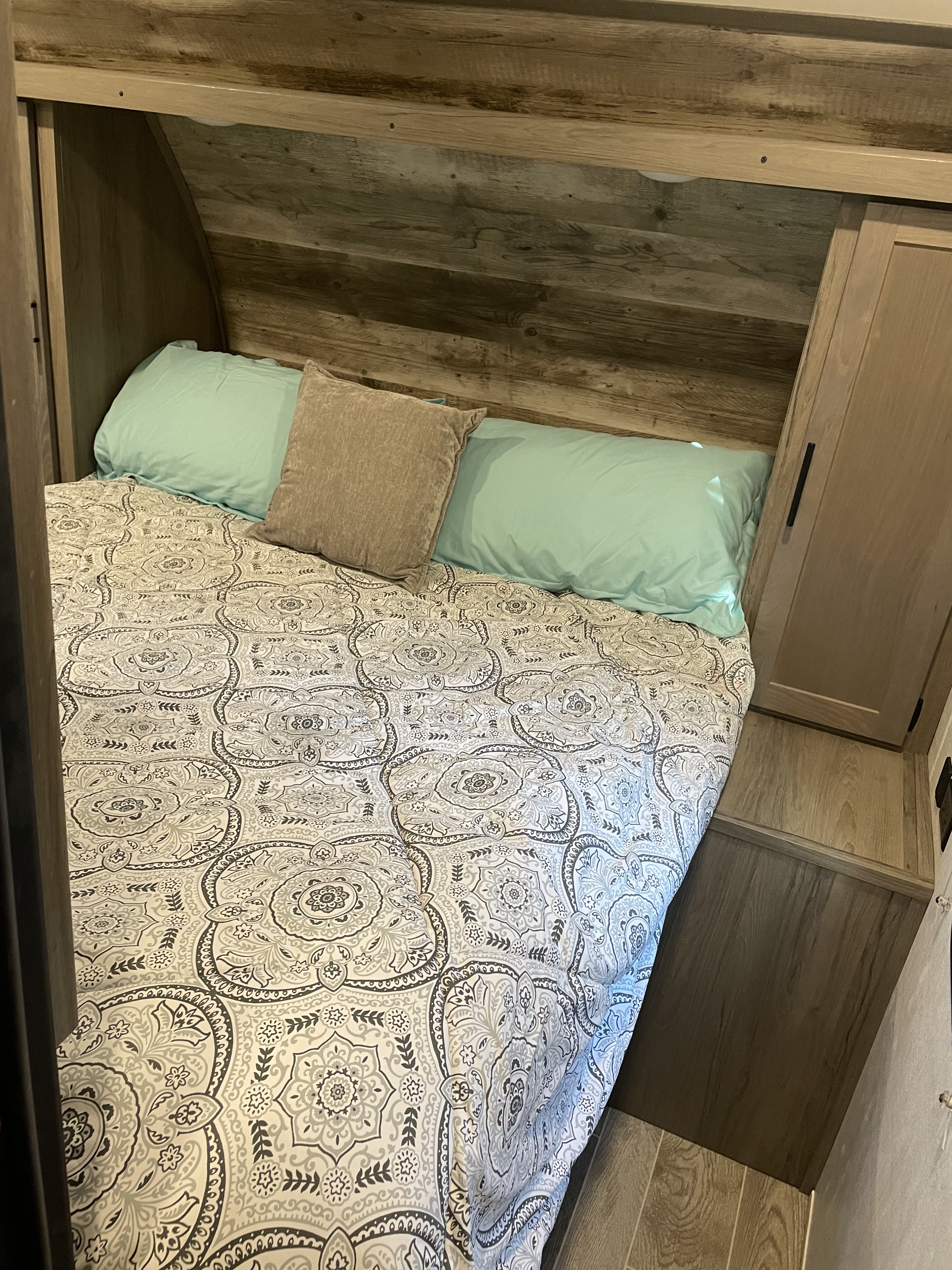 View of an RV rental bedroom featuring a patterned bedspread, light blue and brown pillows, and wooden interior panels.. Gulf Stream Enlighten 2021