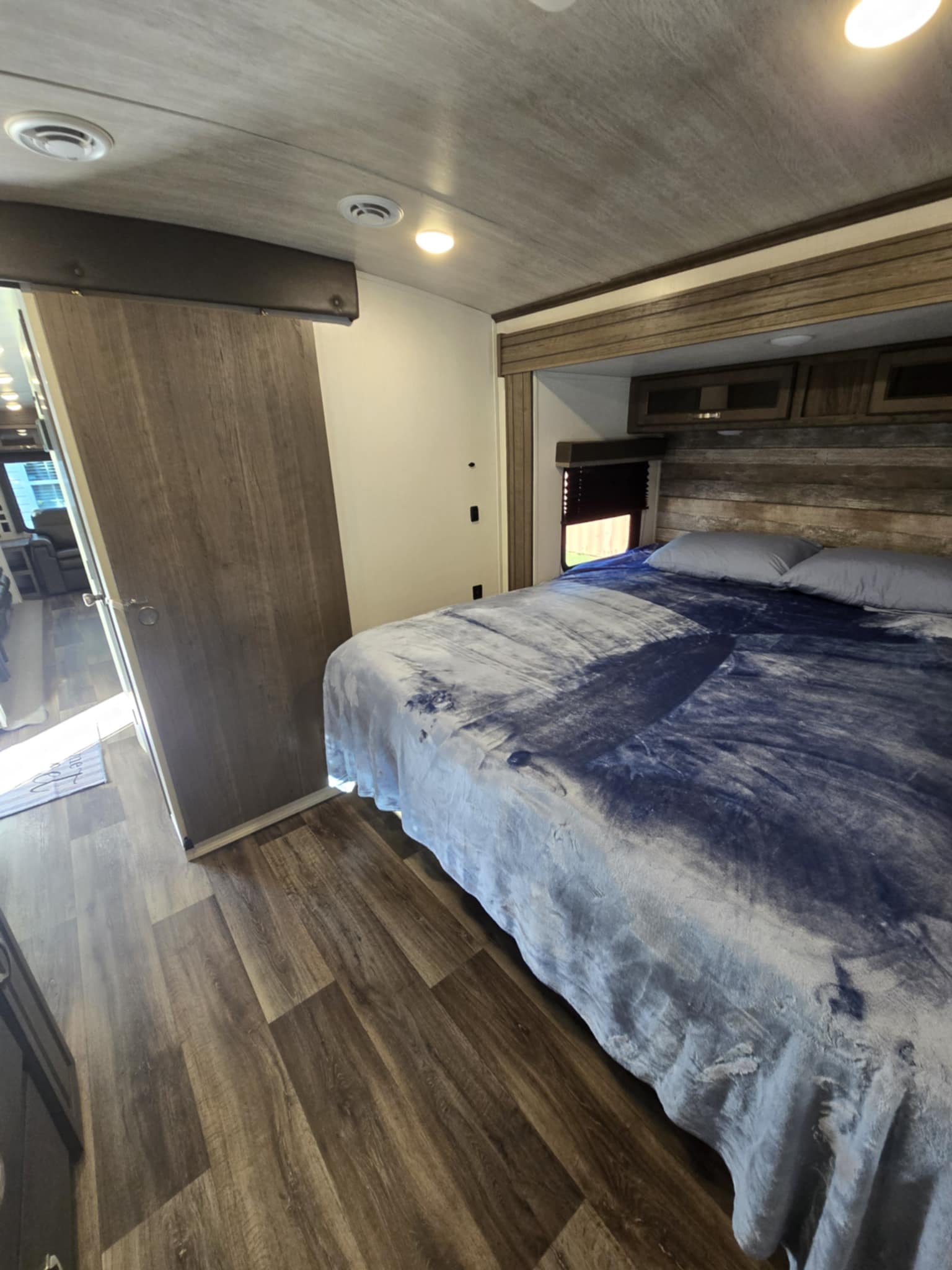 Cozy RV rental bedroom: large bed with blue blanket, wood-look floor, built-in storage, and bright overhead lighting.. Keystone RV Cougar 2021