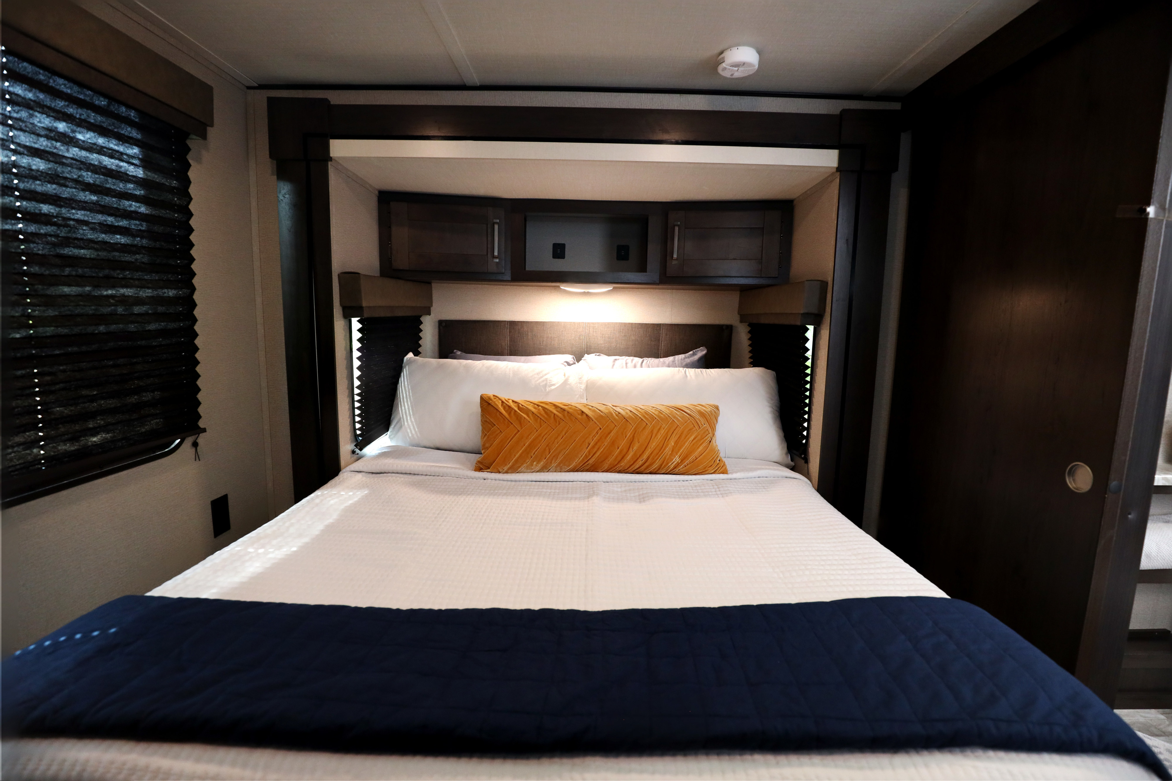 Modern RV bedroom with a cozy queen bed, dark wood accents, lighted headboard, and an orange accent pillow. Ideal for an RV rental.. Grand Design Transcend Xplor 2022