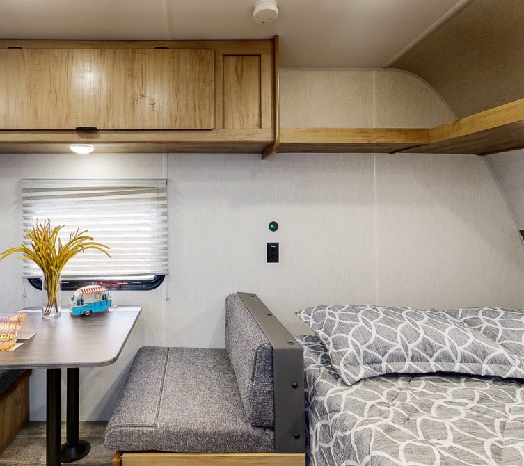 Compact RV rental interior: dining area with window, overhead cabinets, and bed with grey patterned bedding.. Forest River Wolfden 16EV 2024