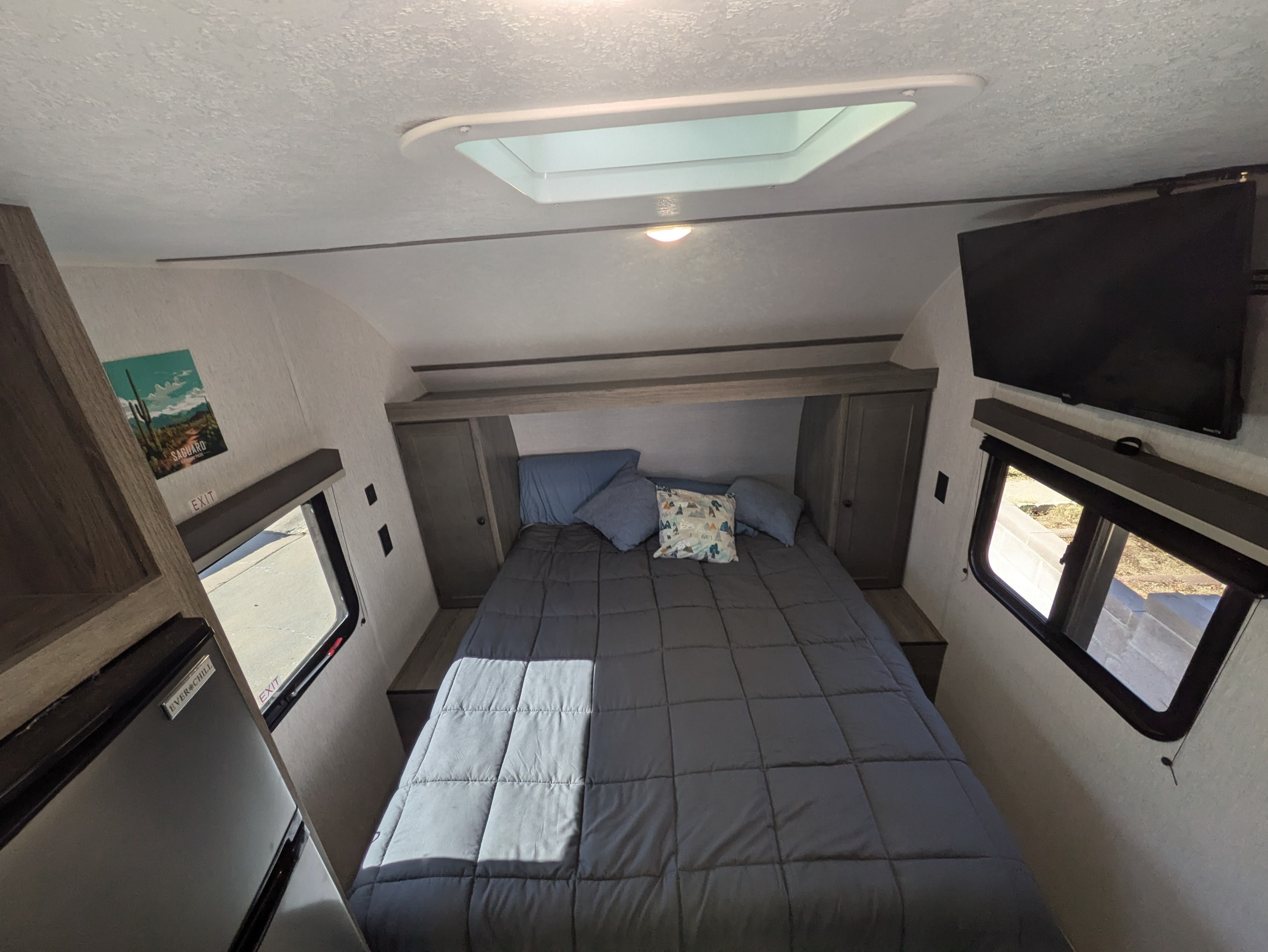 Inside an RV rental, a well-appointed bedroom featuring a large bed, overhead skylight, TV, and fridge.. Sunset Park & Rv Inc. 19rb 2025