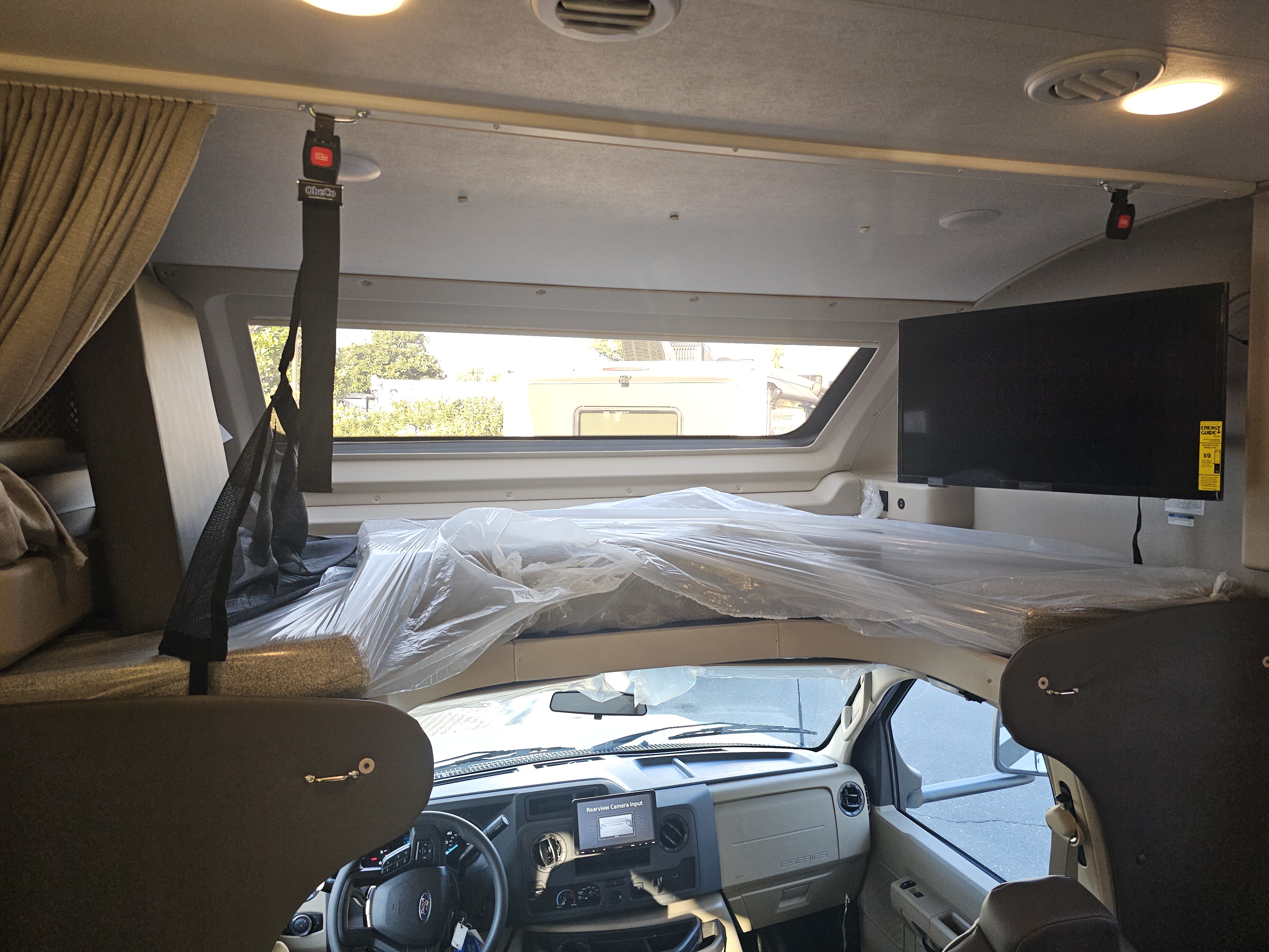 Spacious RV rental cab-over bunk area with safety net, plastic-wrapped mattress, and mounted TV. Front dash visible below.. Jayco Redhawk 2024